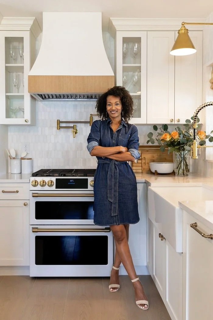 Woman standing in a modern white kitchen with gold accents, wearing a blue denim dress and beige high heels, smiling.
