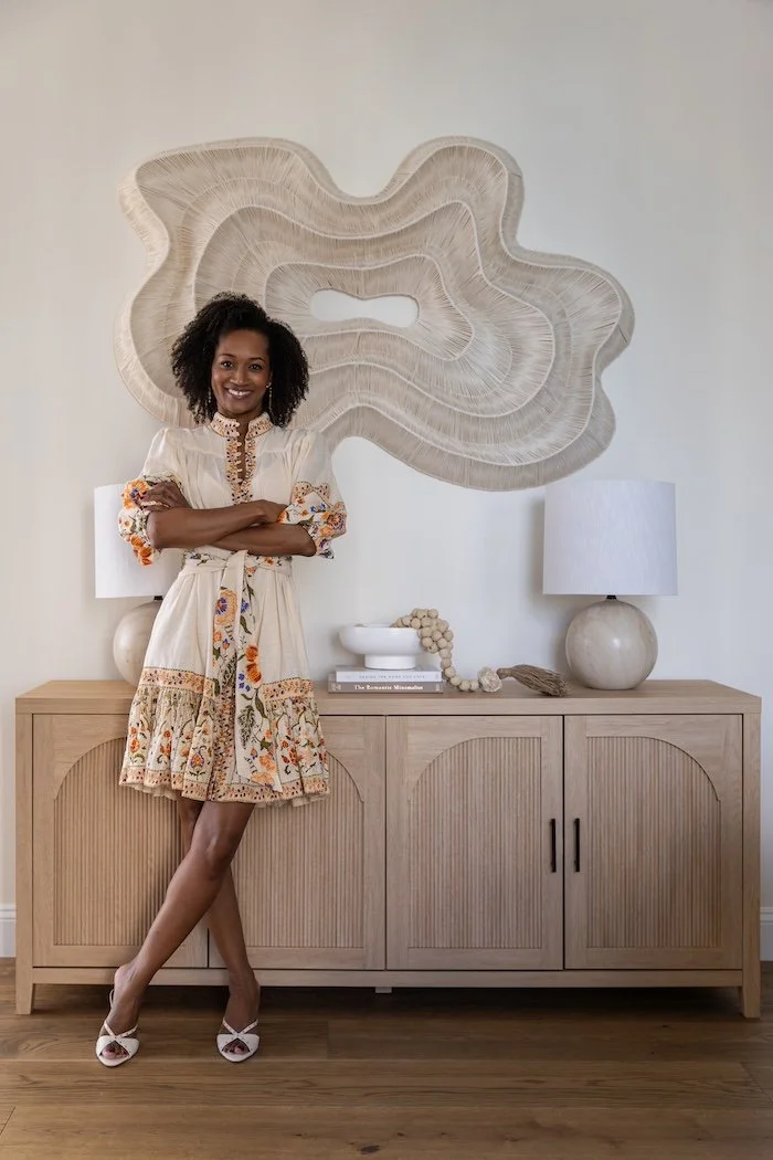 A woman in a cream-colored embroidered dress with floral designs and ruffled sleeves, standing in front of a wooden sideboard with a large, abstract wall art behind her. The sideboard has two white lamps, a white bowl, books, and decorative items. She is smiling with arms crossed.
