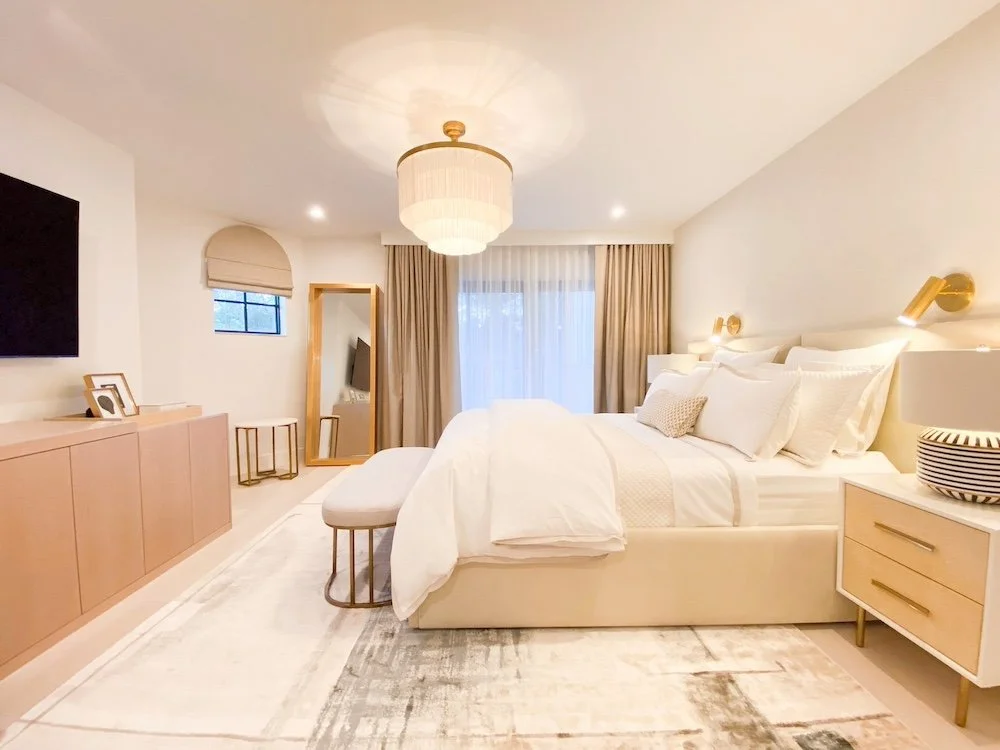 A modern bedroom with a large bed, white linens, and multiple pillows. There are beige curtains covering sliding glass doors, a ceiling light fixture, and a beige and white rug. A dresser with picture frames, a mirror, and a small window is present. Gold wall sconces provide lighting beside the bed. A bench at the foot of the bed and a striped bowl on the nightstand add decorative touches.