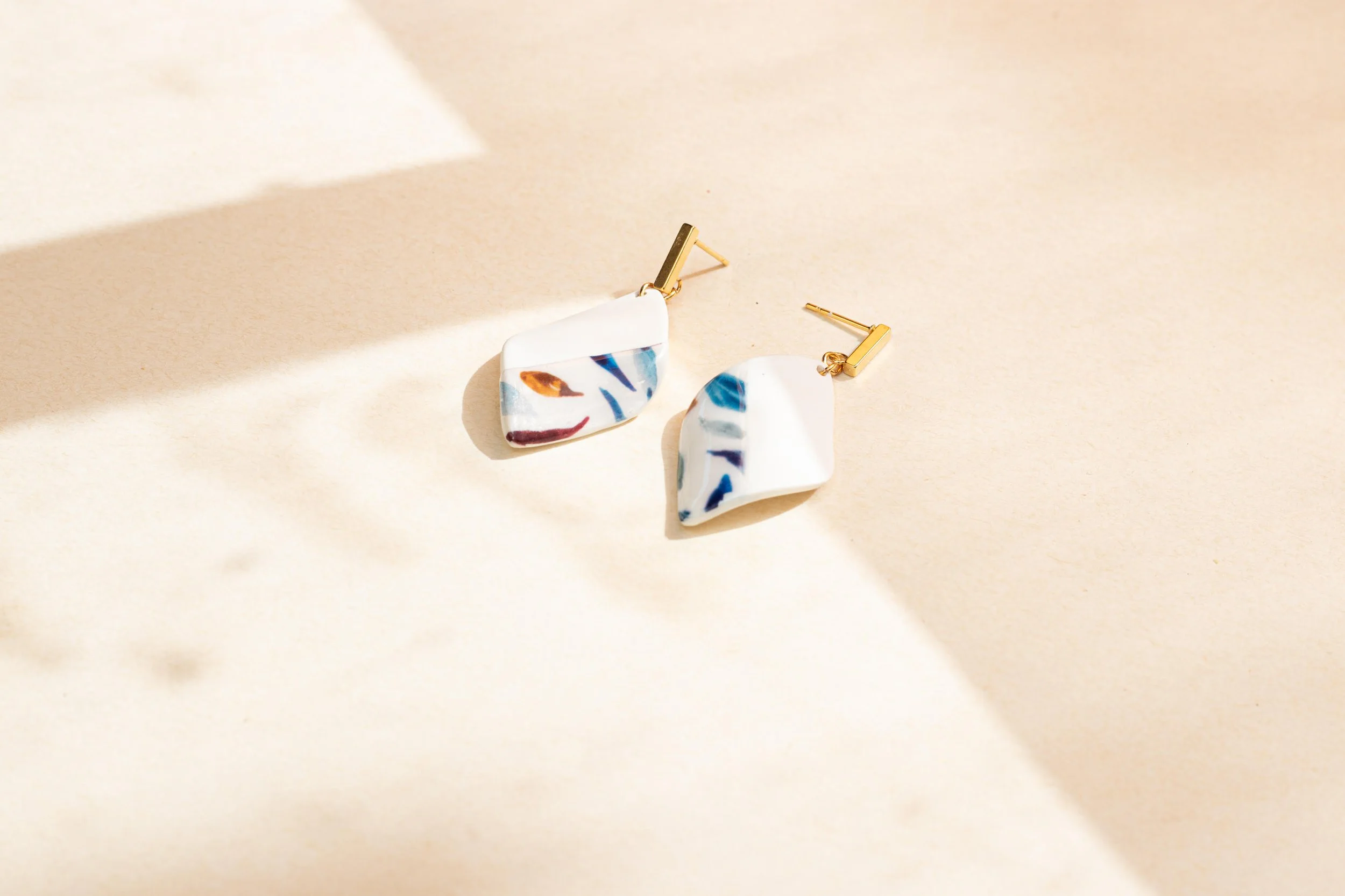 Pair of abstract floral earrings with white, blue, orange, and burgundy colors, laid on a beige surface with shadows.