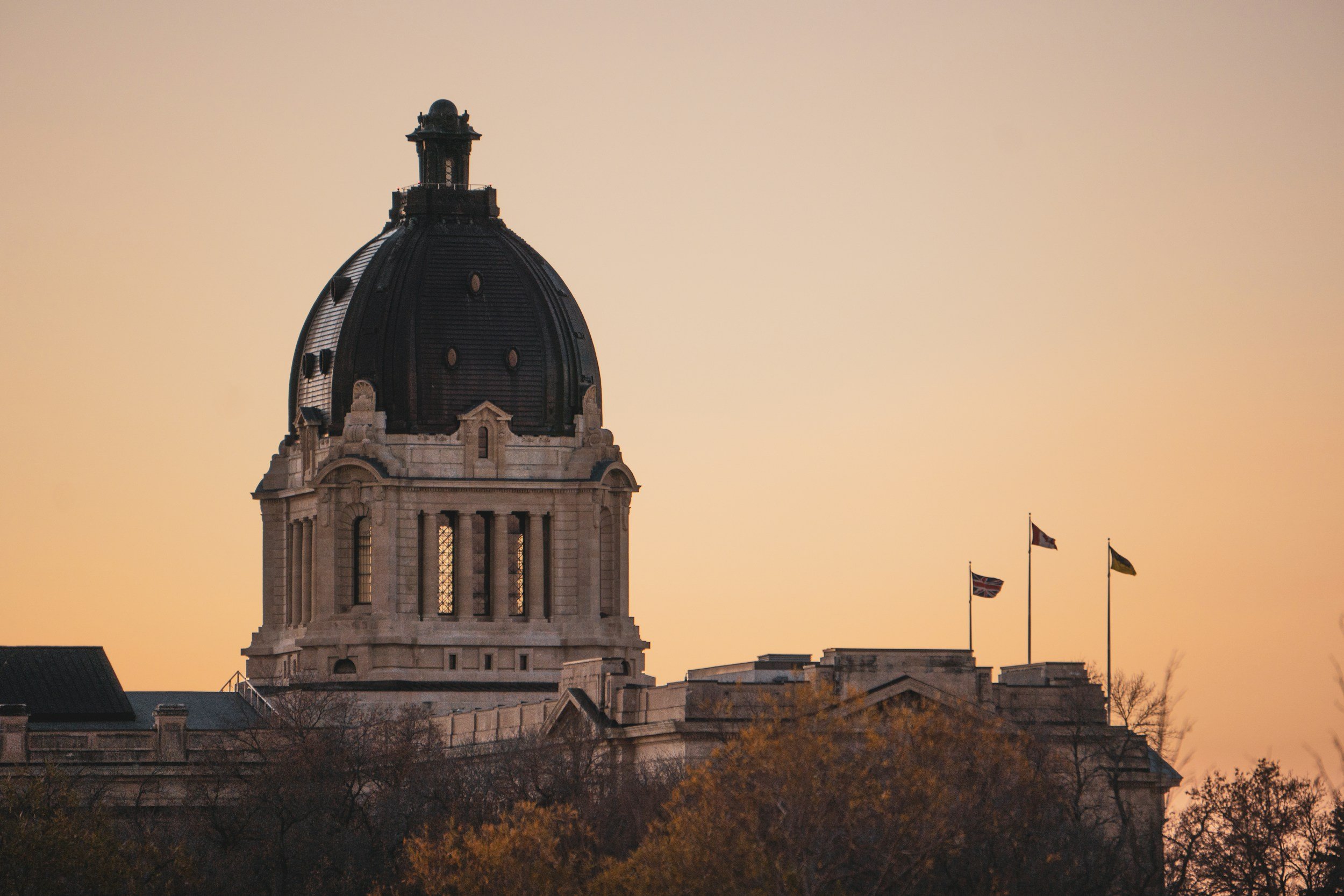 The dome of a grand historic building at sunset, with three flags flying from nearby structures, and trees in the foreground.