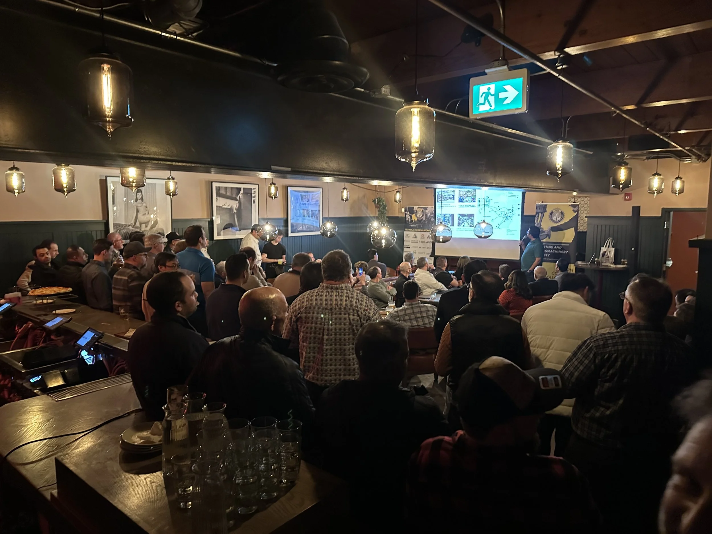 A room filled with people attending a presentation or meeting. There are several large screens displaying information at the front, and a woman is speaking into a microphone. The room is dimly lit with hanging lamps and has a dark ceiling with exposed beams.