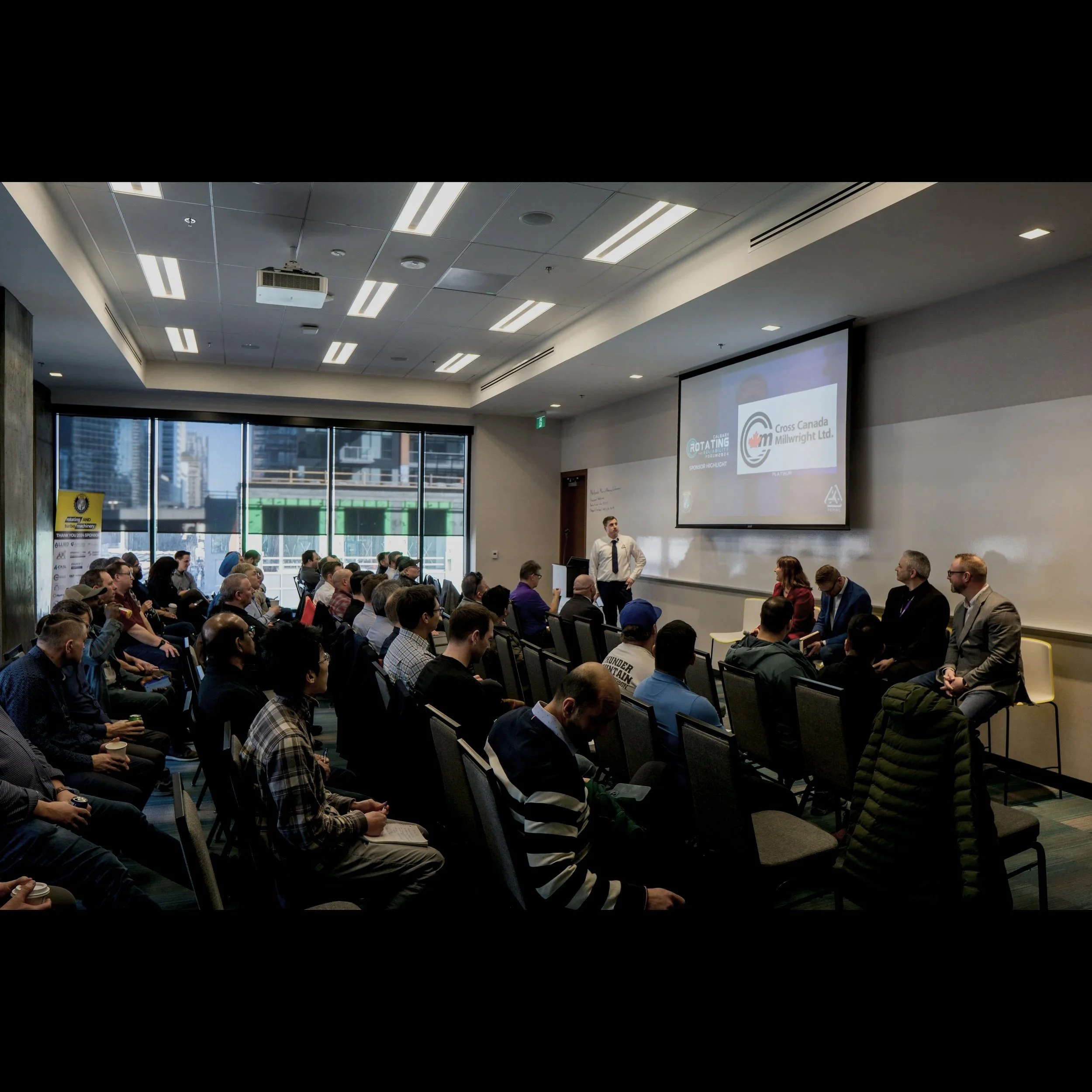 People attending a conference in a modern meeting room with large windows, a presenter speaking at the front, and a screen displaying logos of Cross Canada Millwright Ltd and a rotating sponsorship banner.
