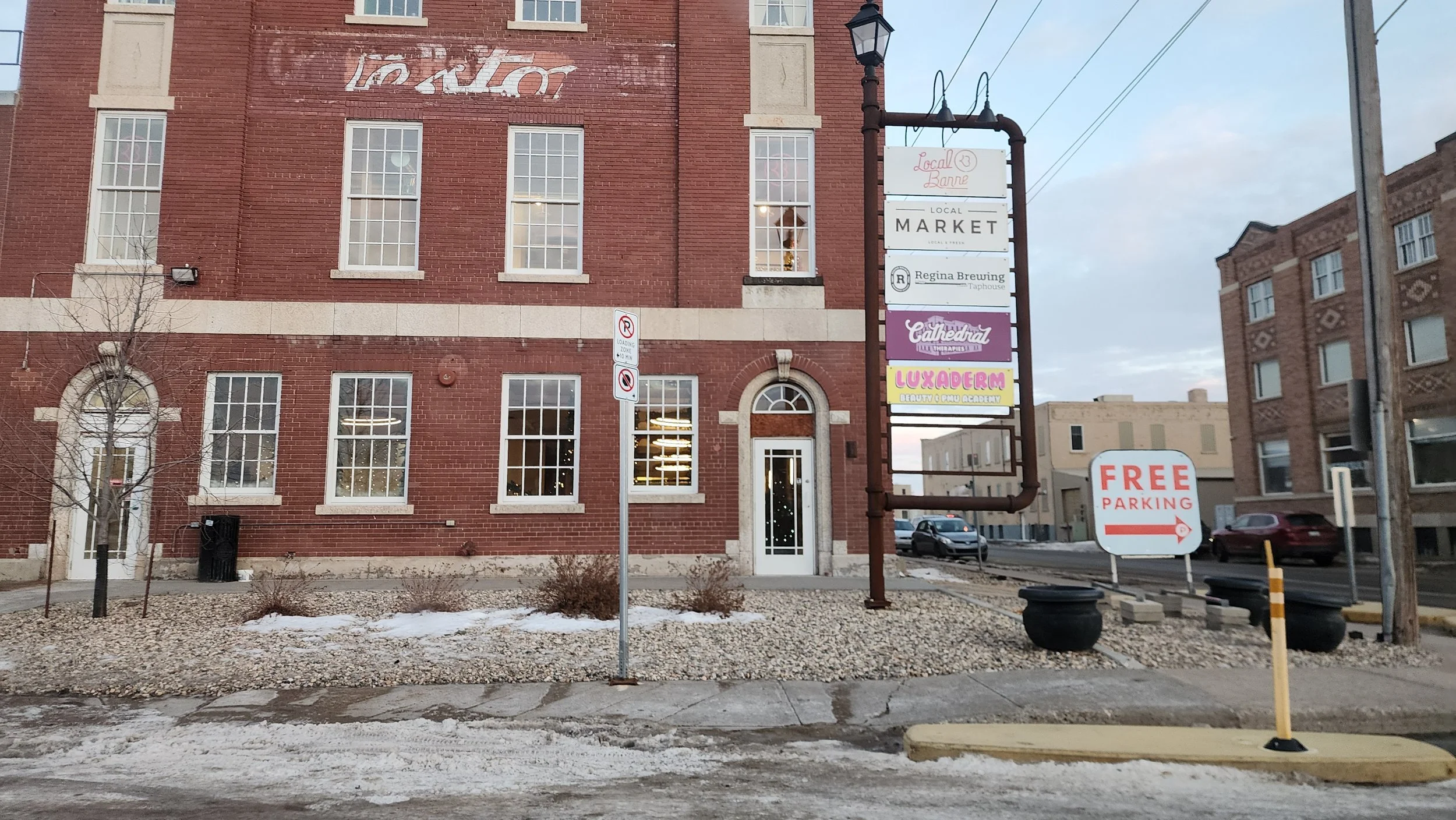 A street scene with a red brick building displaying various business signs, including a local market, Regina Brewing, cathedral therapy, and Luxaderm. There are parking signs, a tree, some snow on the ground, parked cars, and a cloudy sky.