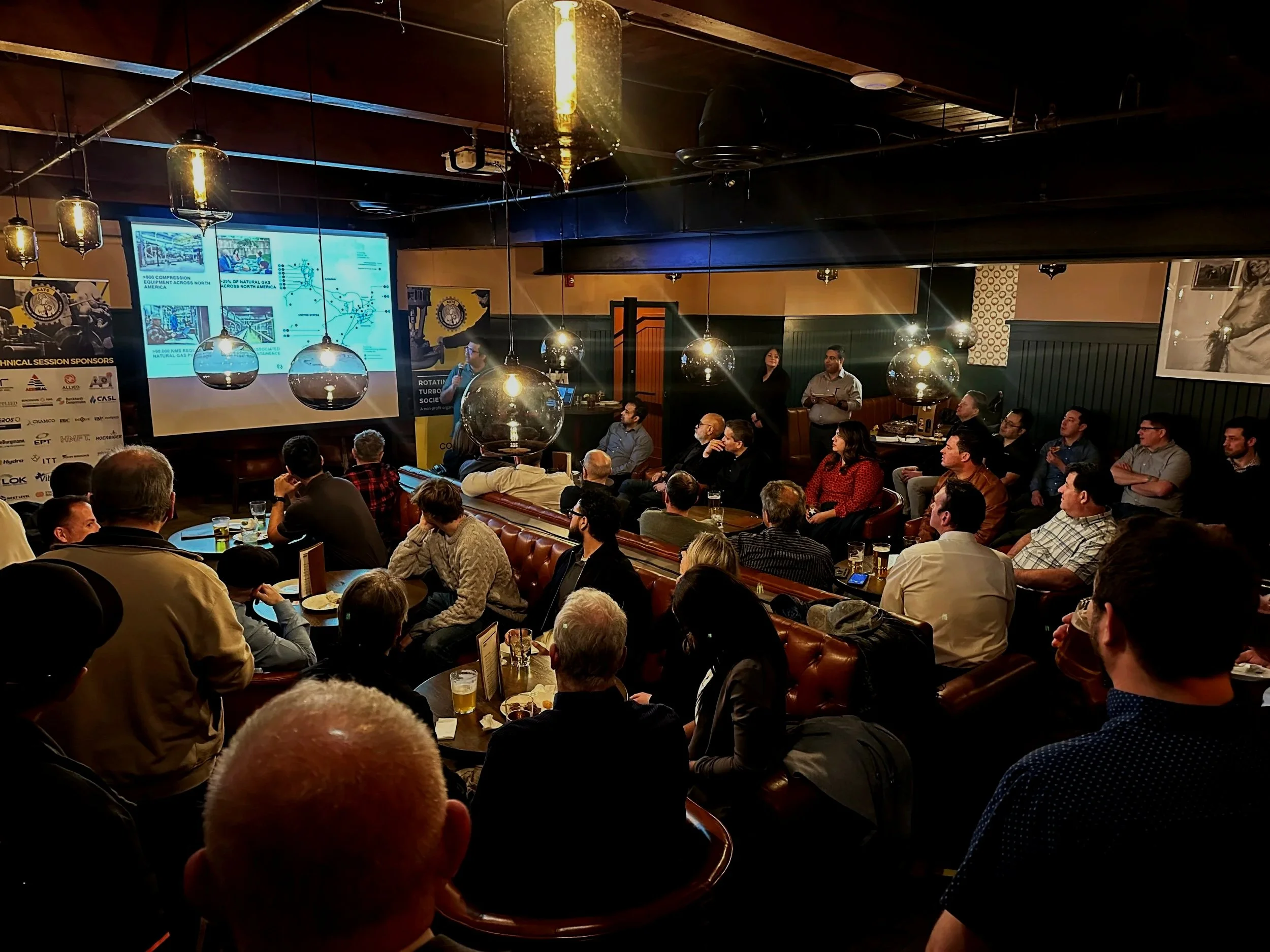 A group of people attending a presentation or conference in a dimly lit room with large screens displaying slides at the front. The audience is seated and focused on the presenter.