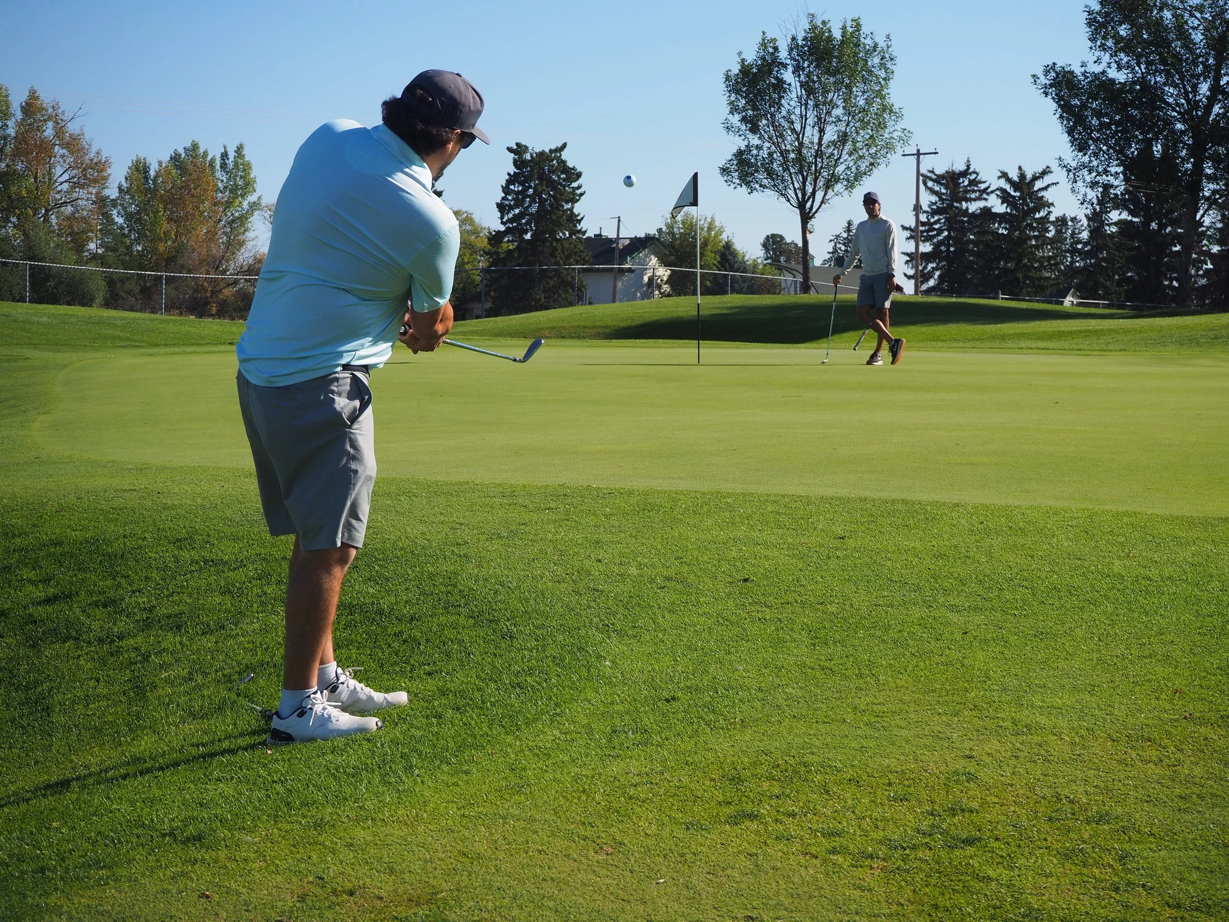 Two men playing golf on a green golf course on a sunny day, one preparing to putt and the other observing.