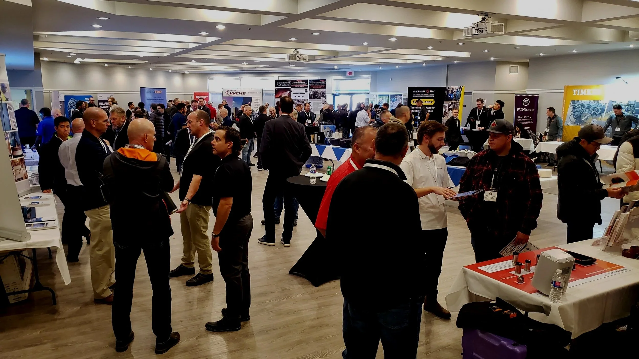 Business professionals at an indoor conference or trade show, walking and talking around exhibitor booths with banners and display tables.