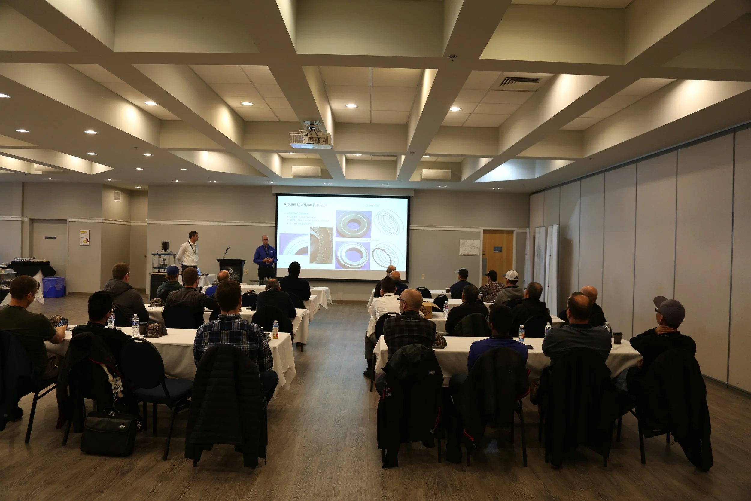 A conference room with attendees sitting at tables listening to a presentation. Two speakers are at the front near a large screen displaying a slide about around the nose gaskets. The room is well-lit with ceiling lights.