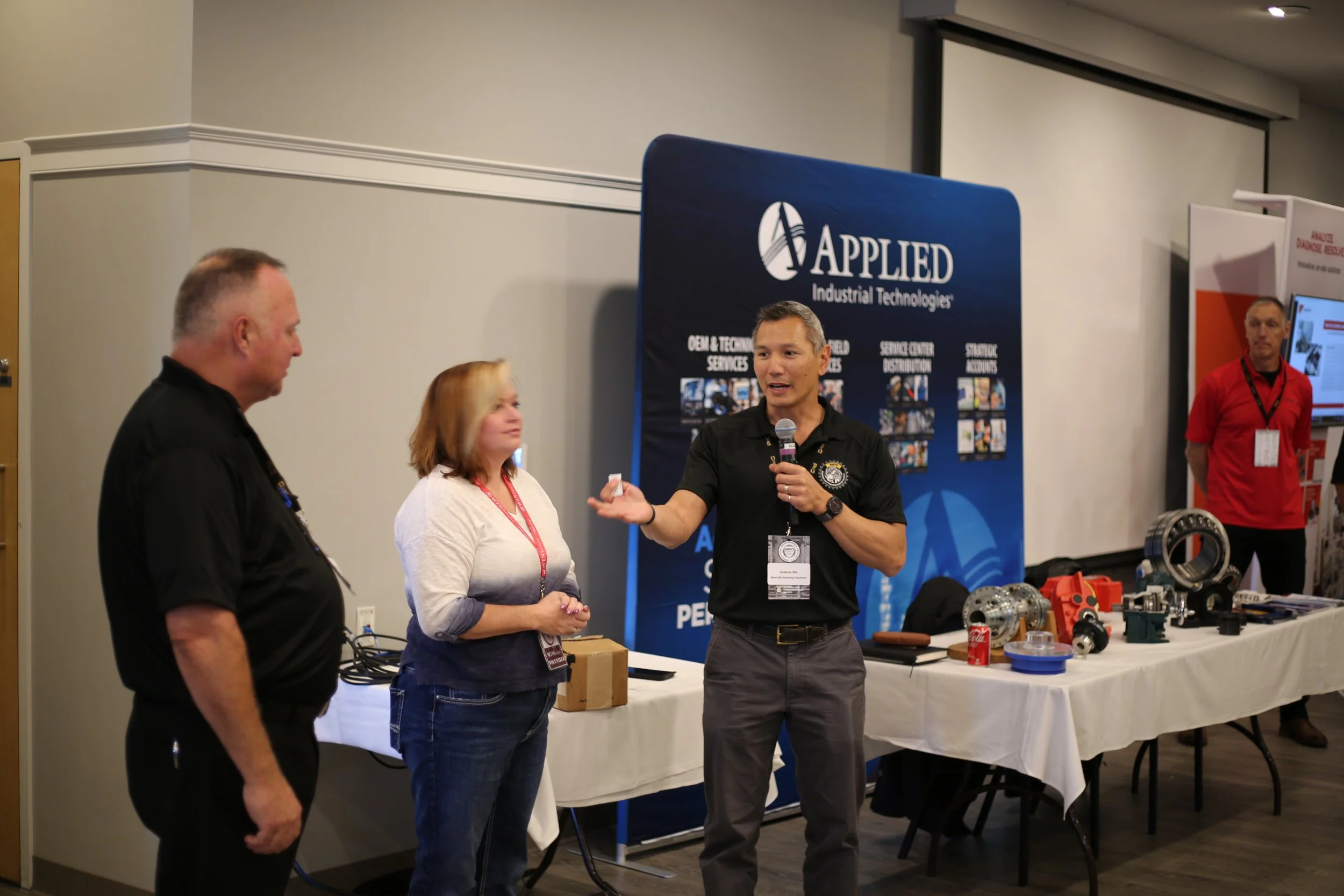 A man giving a presentation at a trade show booth for Applied Industrial Technologies. He is holding a microphone and gesturing to two people standing nearby, a man and a woman, who are listening. The booth has a large blue banner with white text and various industrial parts displayed on a table.