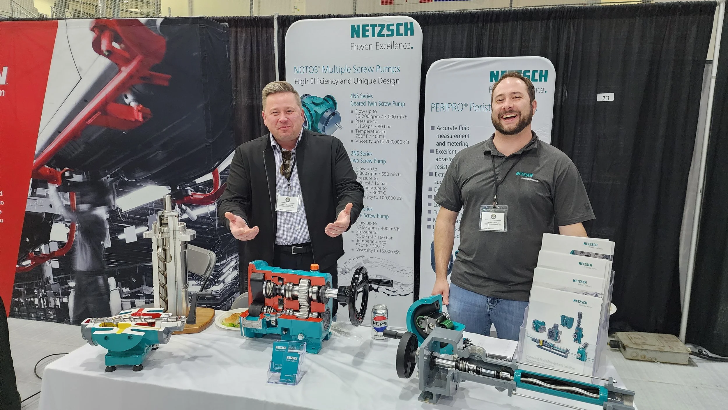 Two men standing behind a table at a trade show booth displaying various industrial pumps and mechanical parts, with informational posters and brochures in the background.