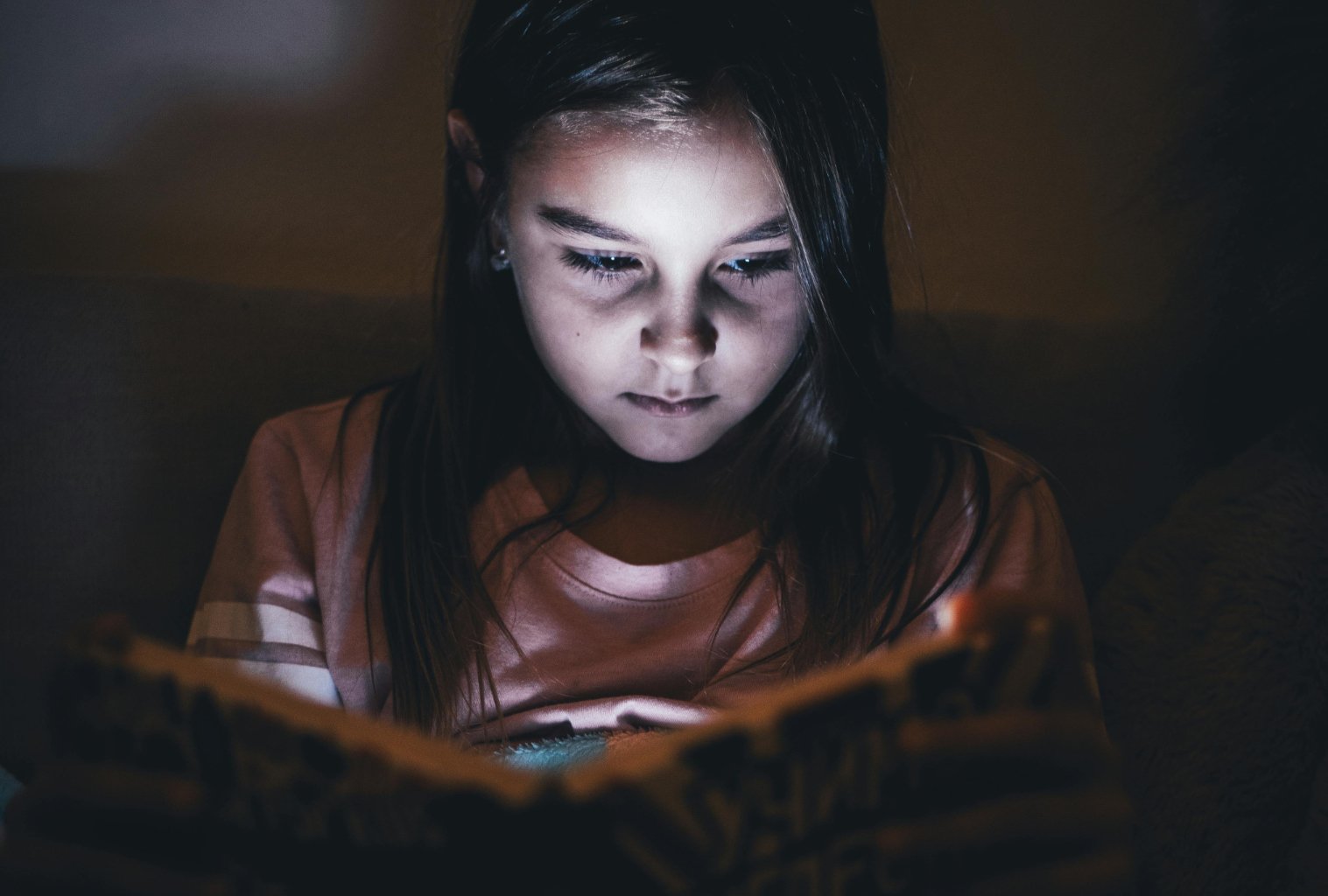 **ALT text:** Young girl sitting in a dimly lit room, her face softly illuminated by the glow of a book or screen she is reading, conveying deep focus, curiosity, and quiet immersion in a story.