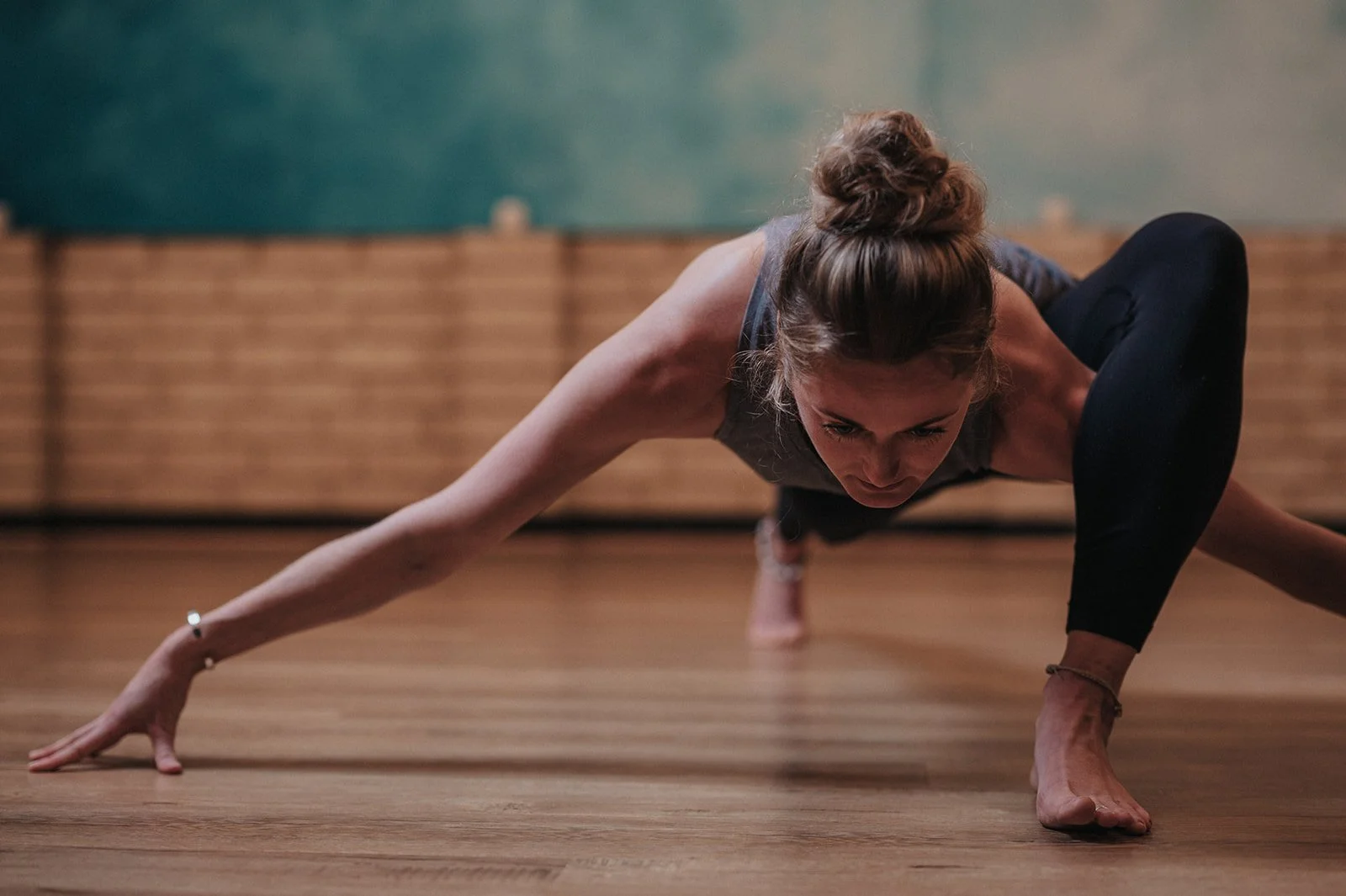 A woman practicing yoga or stretching on a wooden floor in a gym or studio, in a wide low lunge position with arms extended and head lowered.