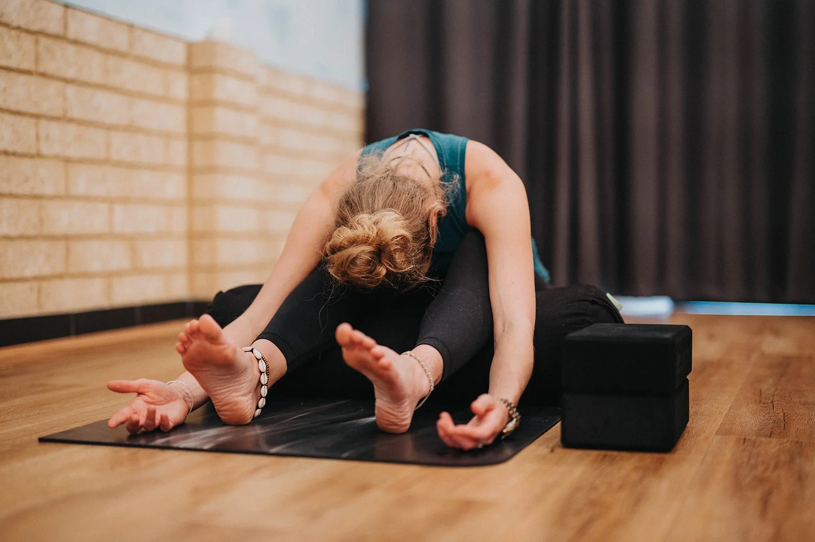 A woman practicing yoga on a black mat with another person, lying on the mat. They are in a stretching pose with the woman leaning forward over the person, who is lying with legs bent and feet on the ground, wearing black leggings and a blue top. The setting appears to be a yoga studio with wooden floor, brick wall, and dark curtain.