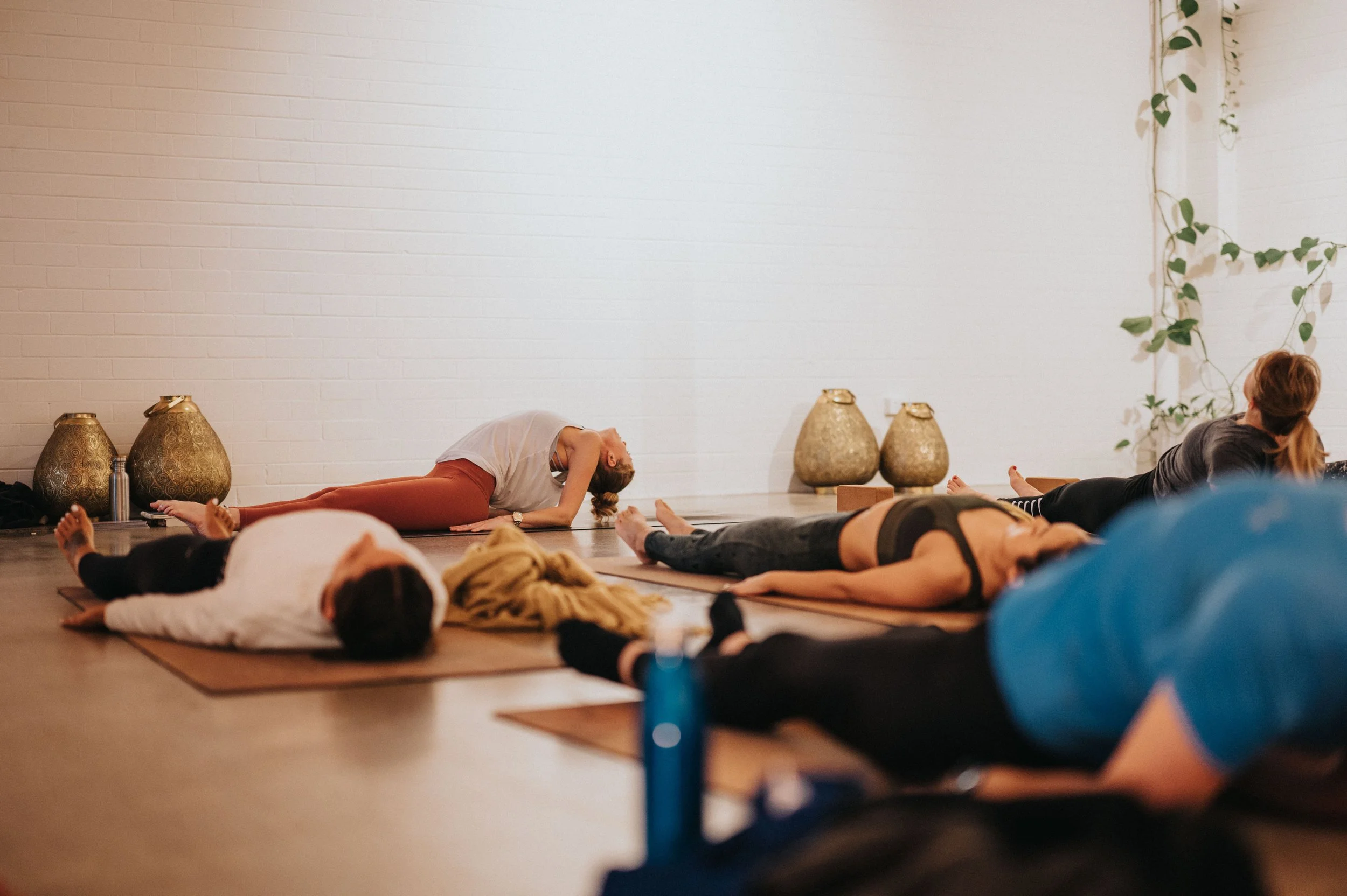 People participating in a yoga class lying on mats in a serene studio with a white brick wall and decorative vases.