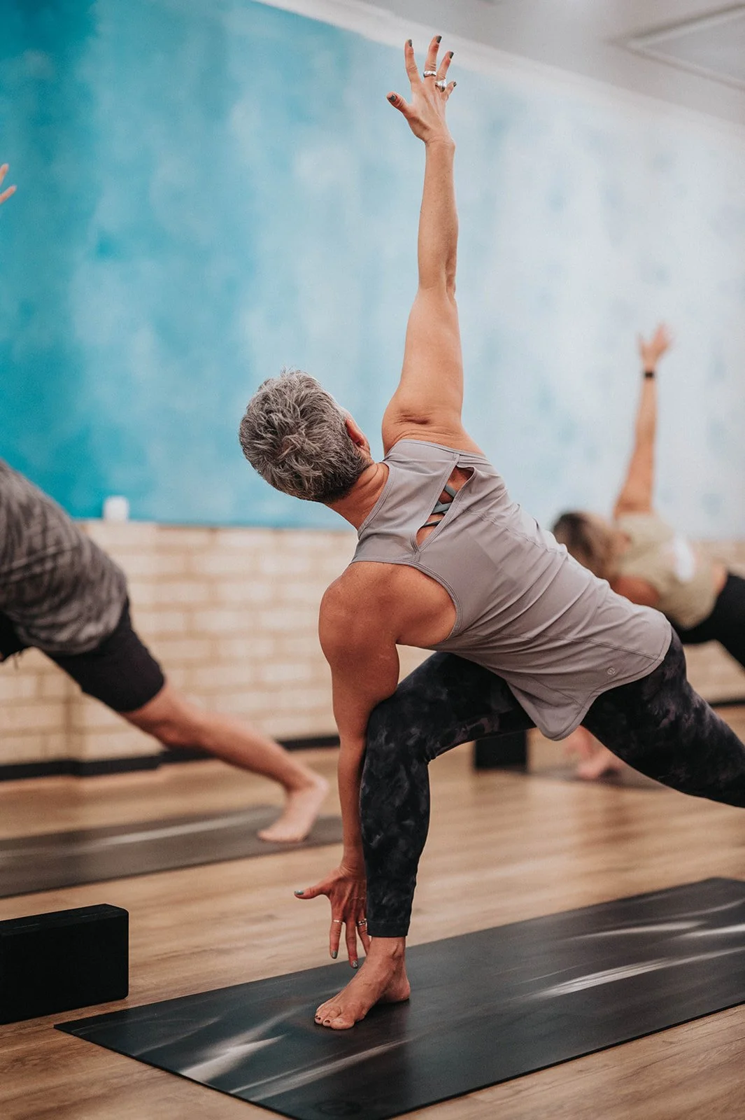 Older woman practicing yoga in a lunge pose with one arm raised in a yoga studio, other participants in the background.