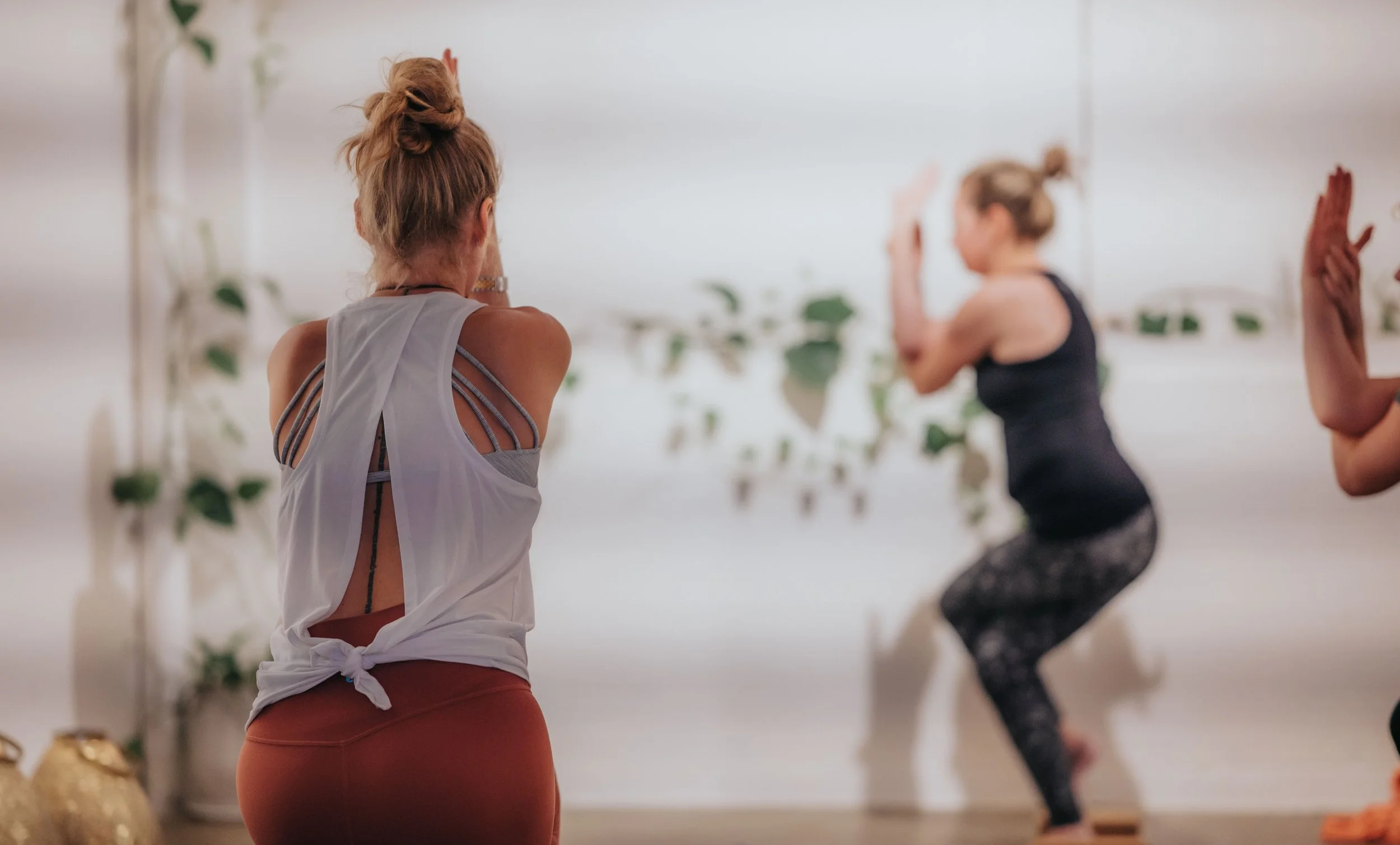 Women practicing yoga in a studio, with one woman in the foreground facing away and others in the background performing poses.