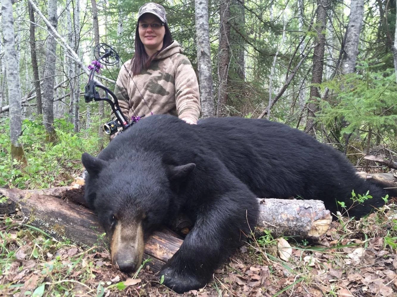 A woman in camouflage and glasses sitting in a forest, holding a bow, next to a large black bear lying on the ground.