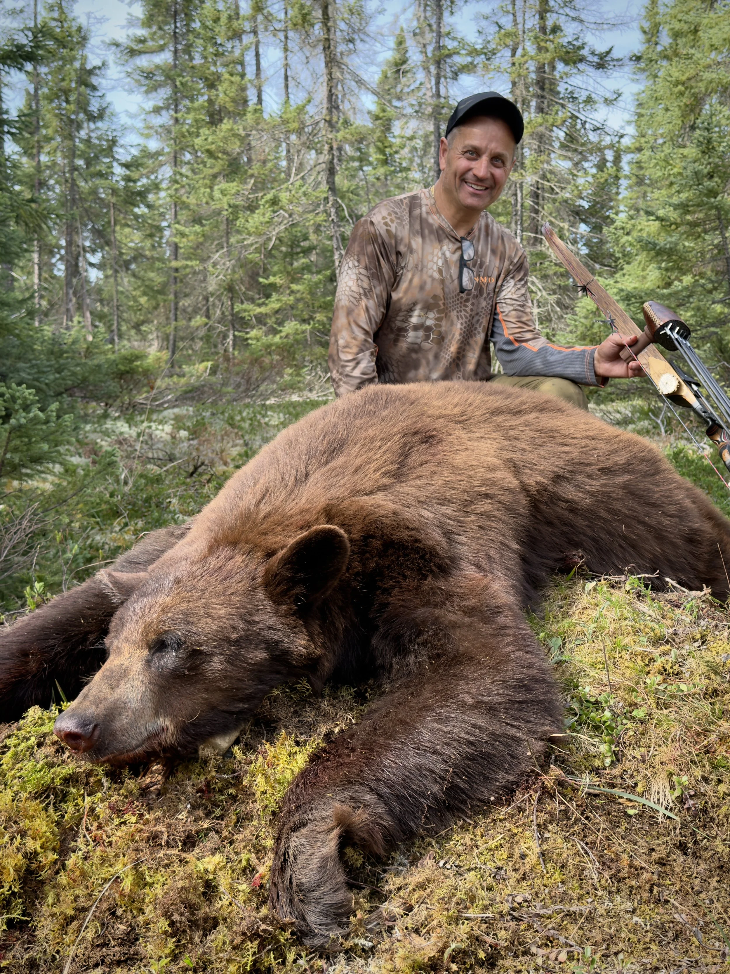A man in camouflage clothing smiles as he holds a bow, kneeling next to a large black bear lying on the ground in a forest of tall green trees.