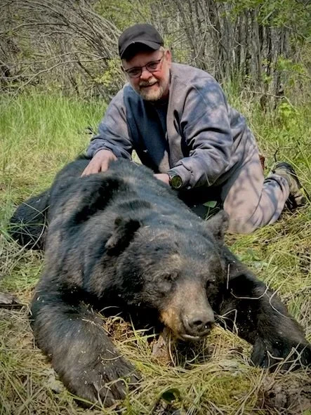 A man kneeling next to a large black bear in a forested area.
