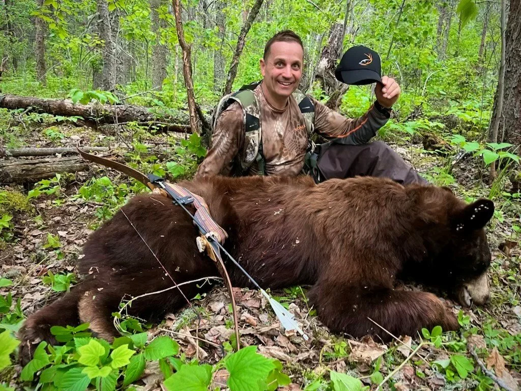 A man in camouflage is sitting in a forest with a large brown bear. He is smiling and holding a hat with the Target Species Outfitting logo, with a bow resting against the bear.