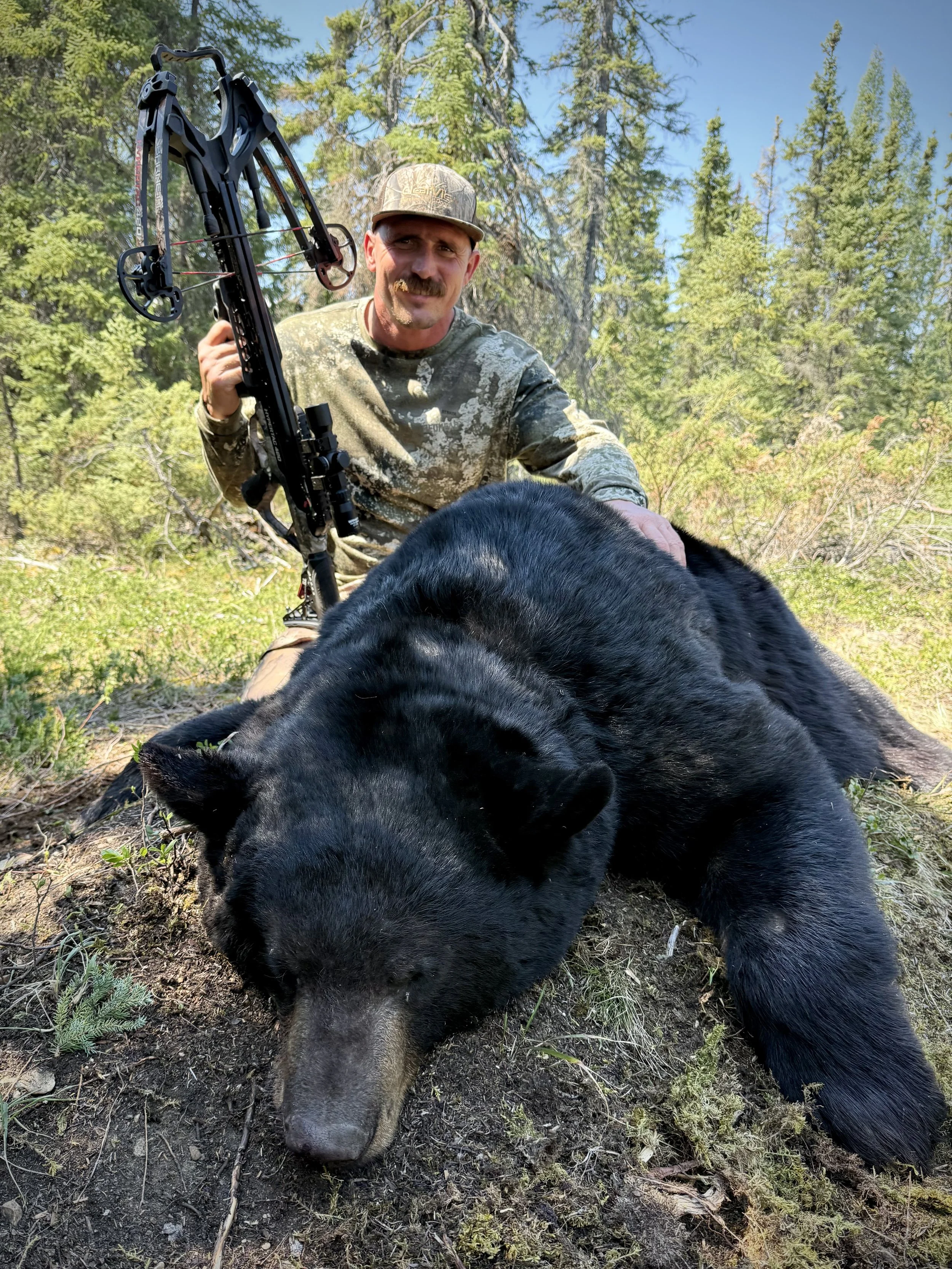 A man in camouflage clothing and a baseball hat is kneeling in a forest next to a black bear, holding a crossbow.
