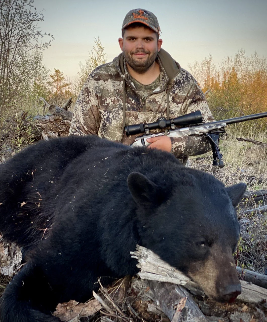 A man with a beard and camouflage kneels in a field, holding a rifle with a scope, next to a black bear that is lying on the ground.