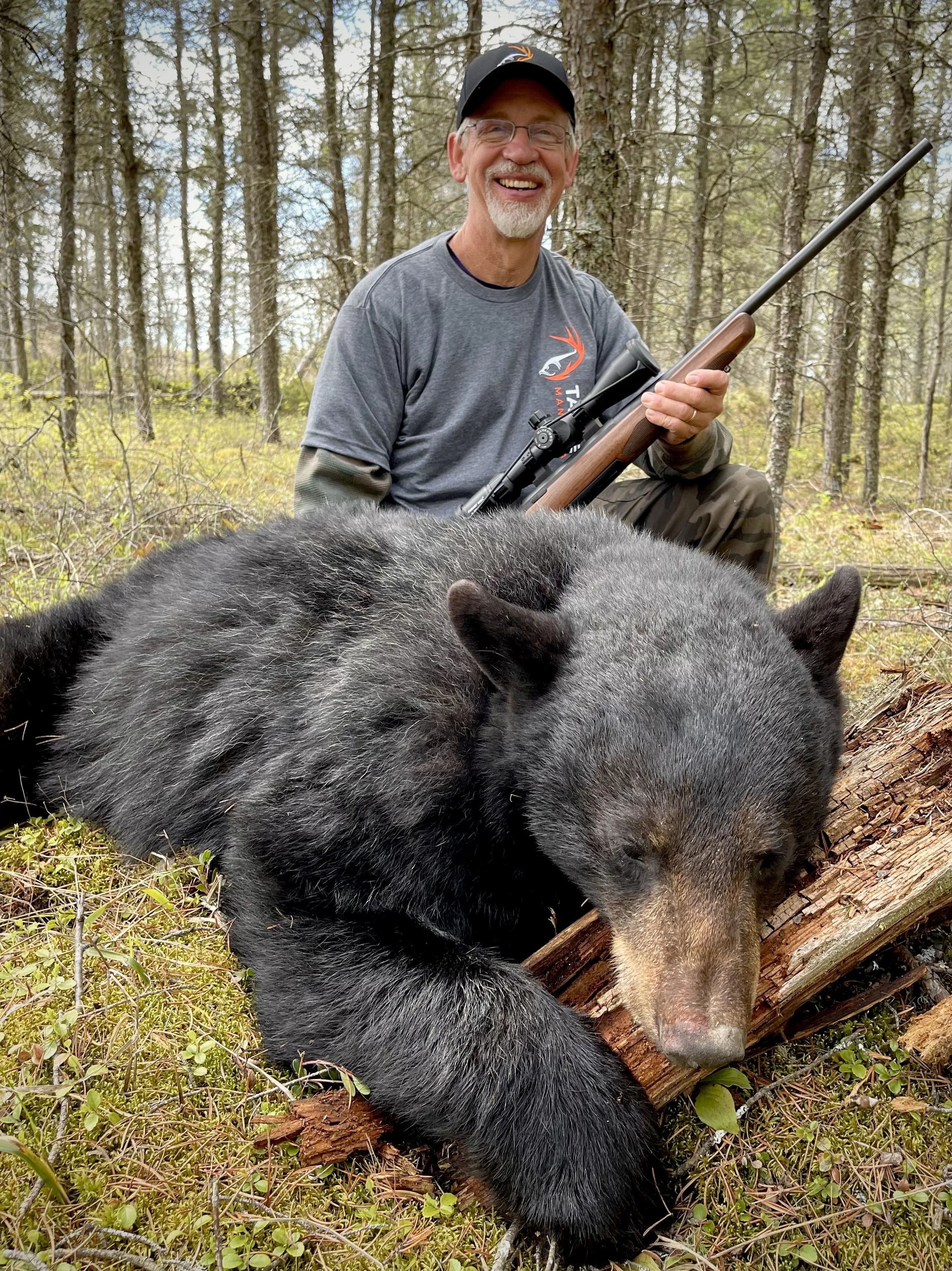 A man in outdoor clothing holding a scoped rifle and smiling, next to a large black bear lying on a fallen log in a forested area.