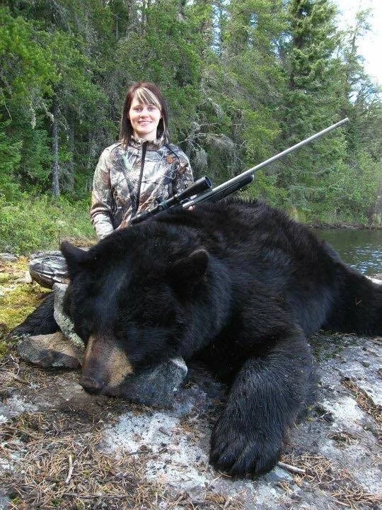A woman in camouflage is kneeling next to a large, black bear lying on the ground near a river, holding a rifle. 