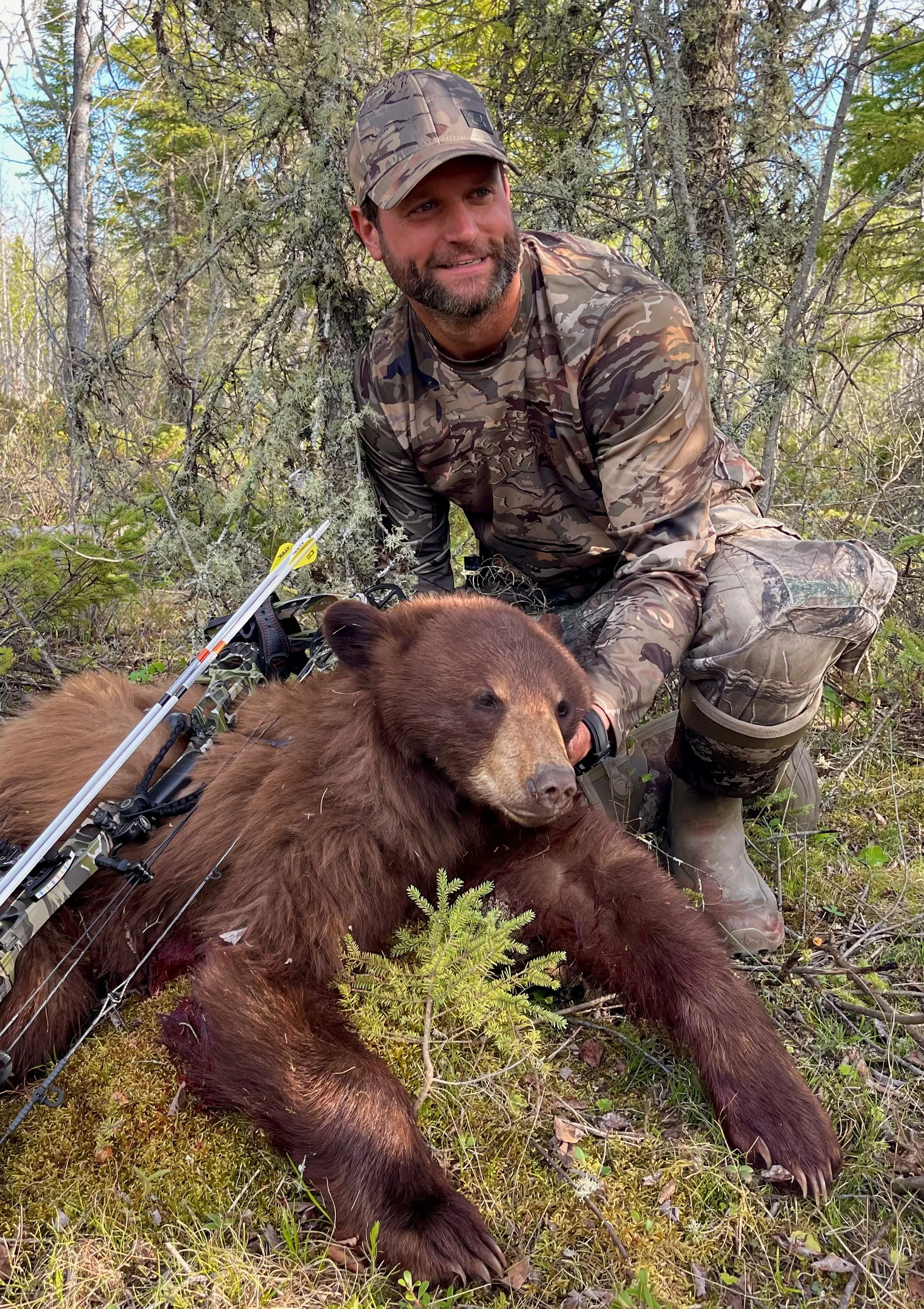 A man in camouflage kneels next to a brown bear in a forested area with a bow lying on the ground.