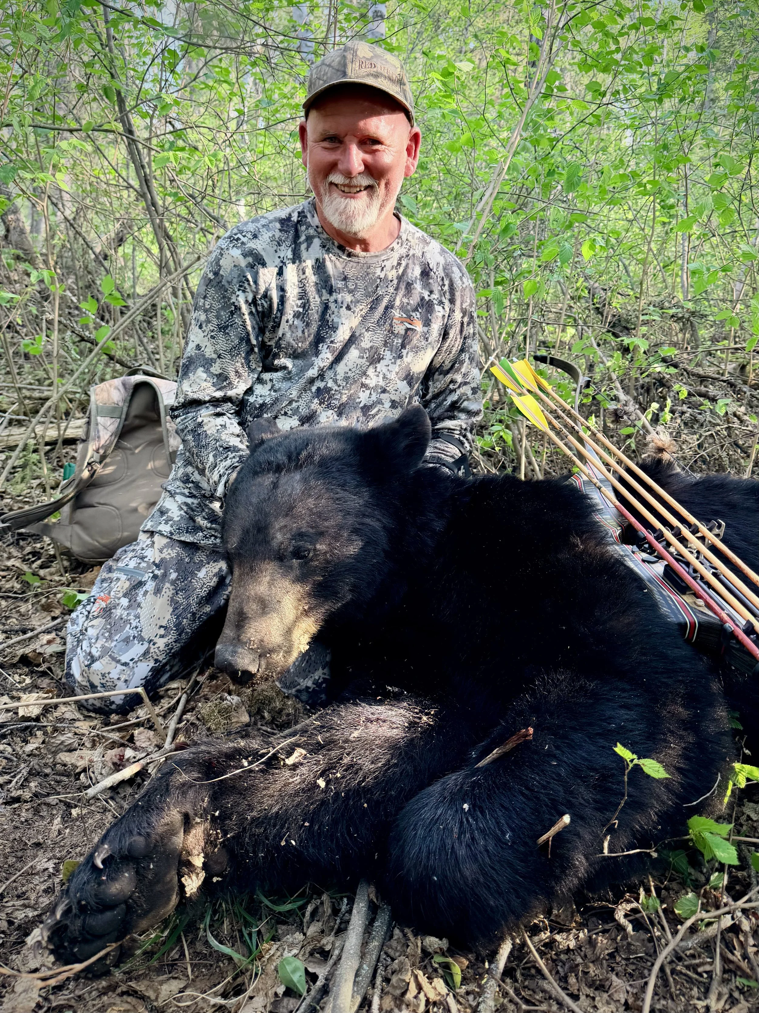 A smiling man in camouflage and a baseball hat kneeling in a forest, with a black bear and arrows on its back.