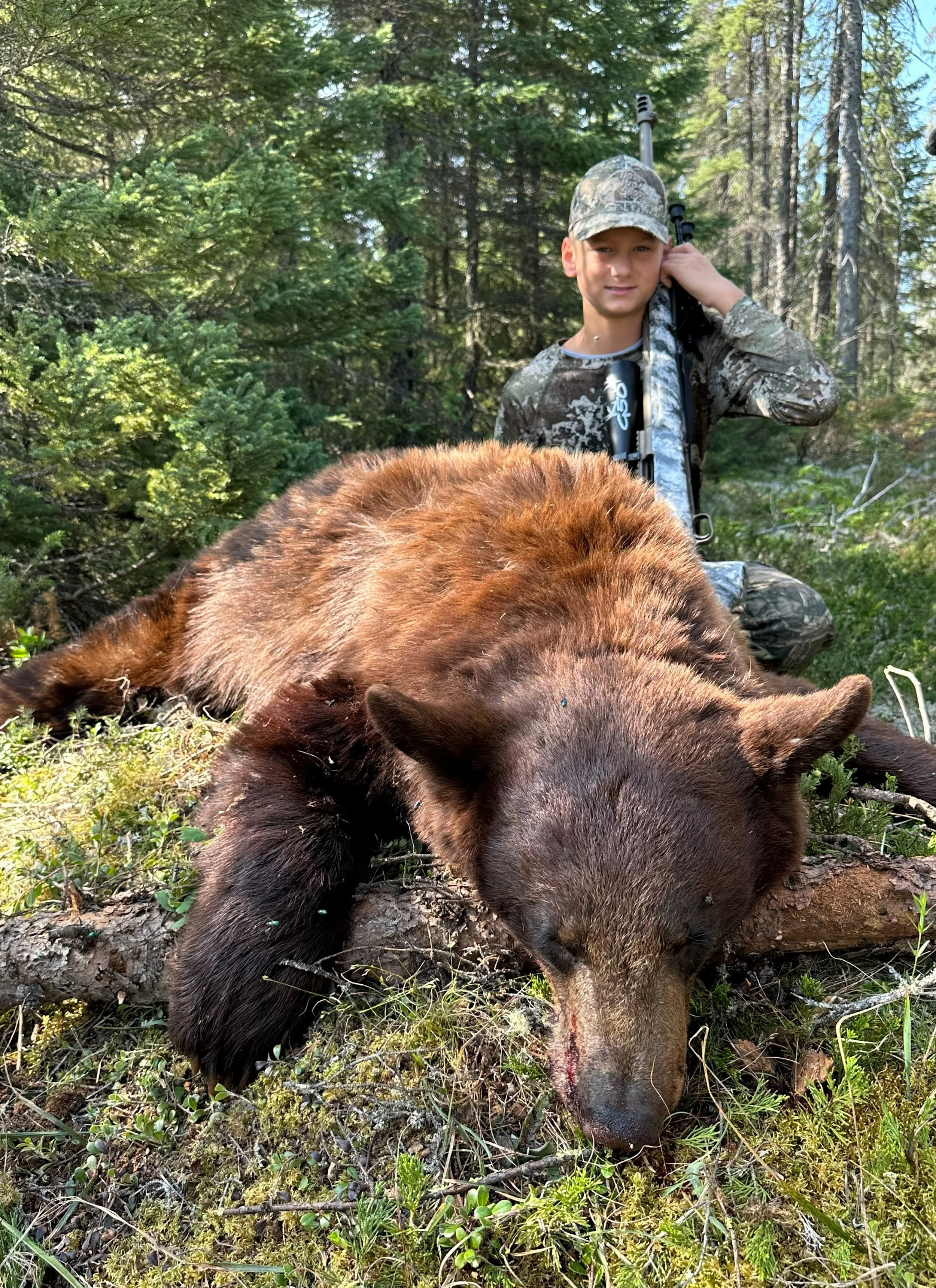 A boy in camouflage clothing holds a rifle behind a brown bear lying on the forest ground surrounded by green trees and foliage.