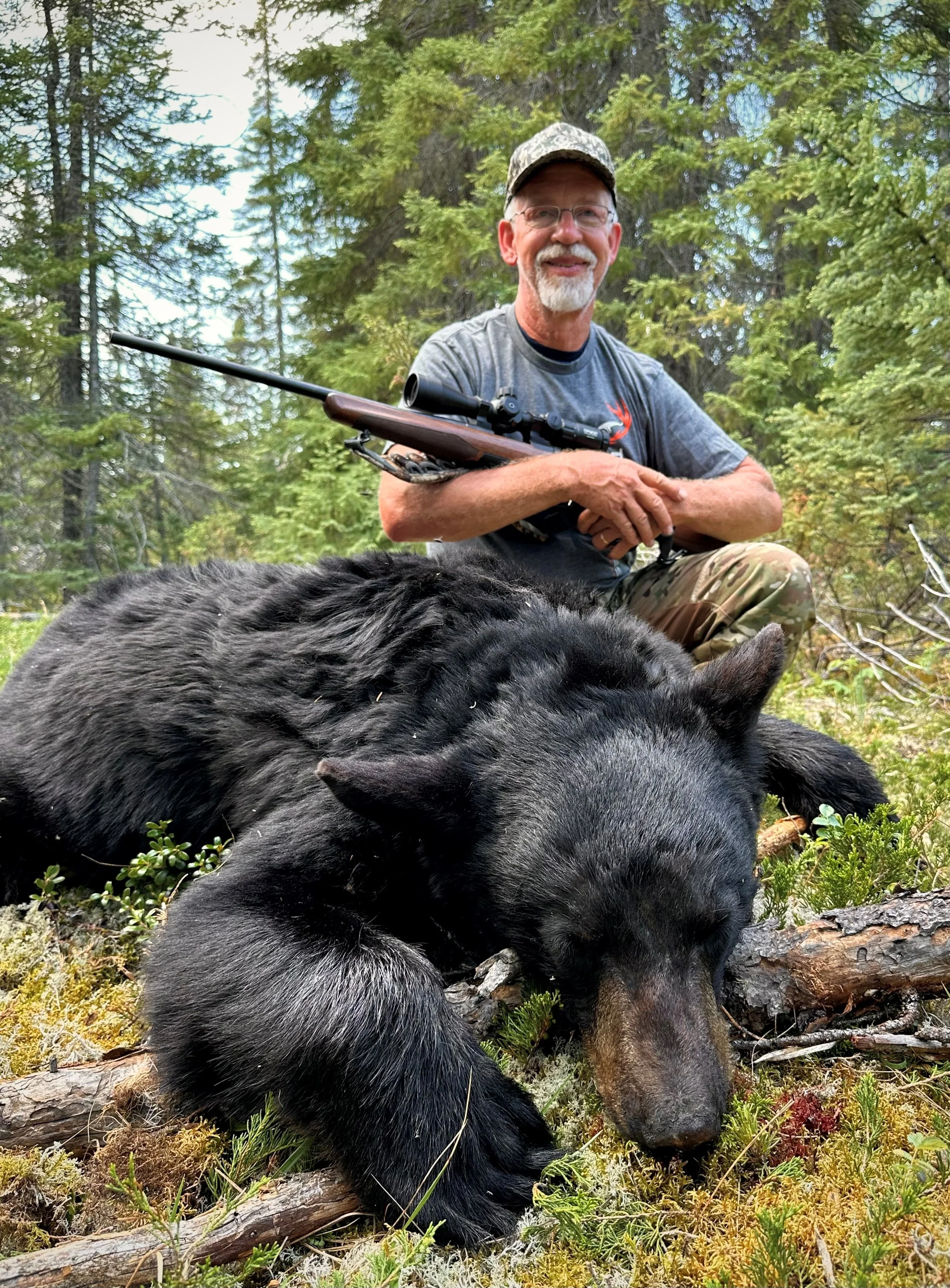 A man kneeling in a forest with a rifle, smiling, with a large black bear lying on the ground.