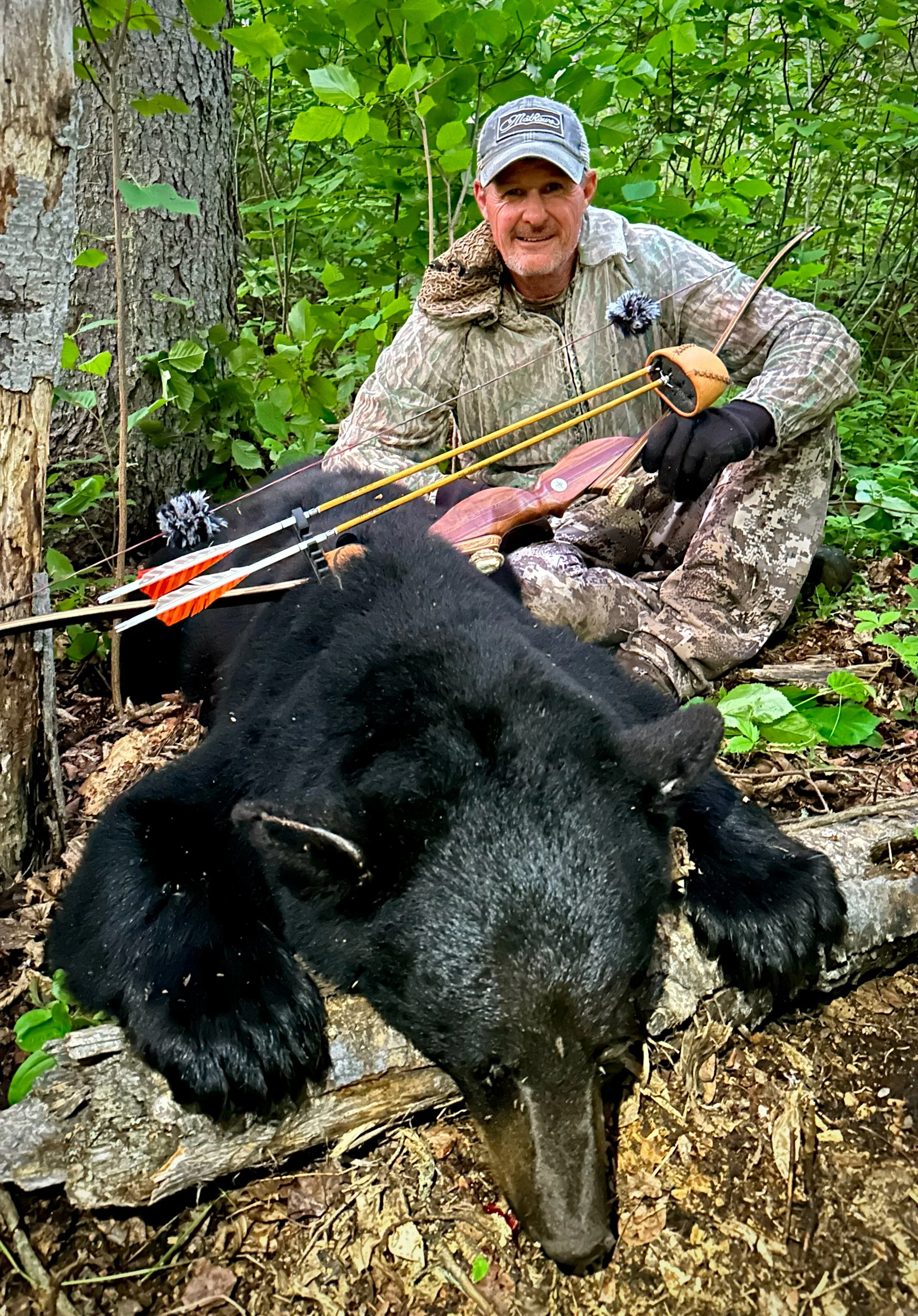 A smiling man in camouflage and a gray hat is sitting in the woods next to a black bear, with a bow and arrow lying nearby.