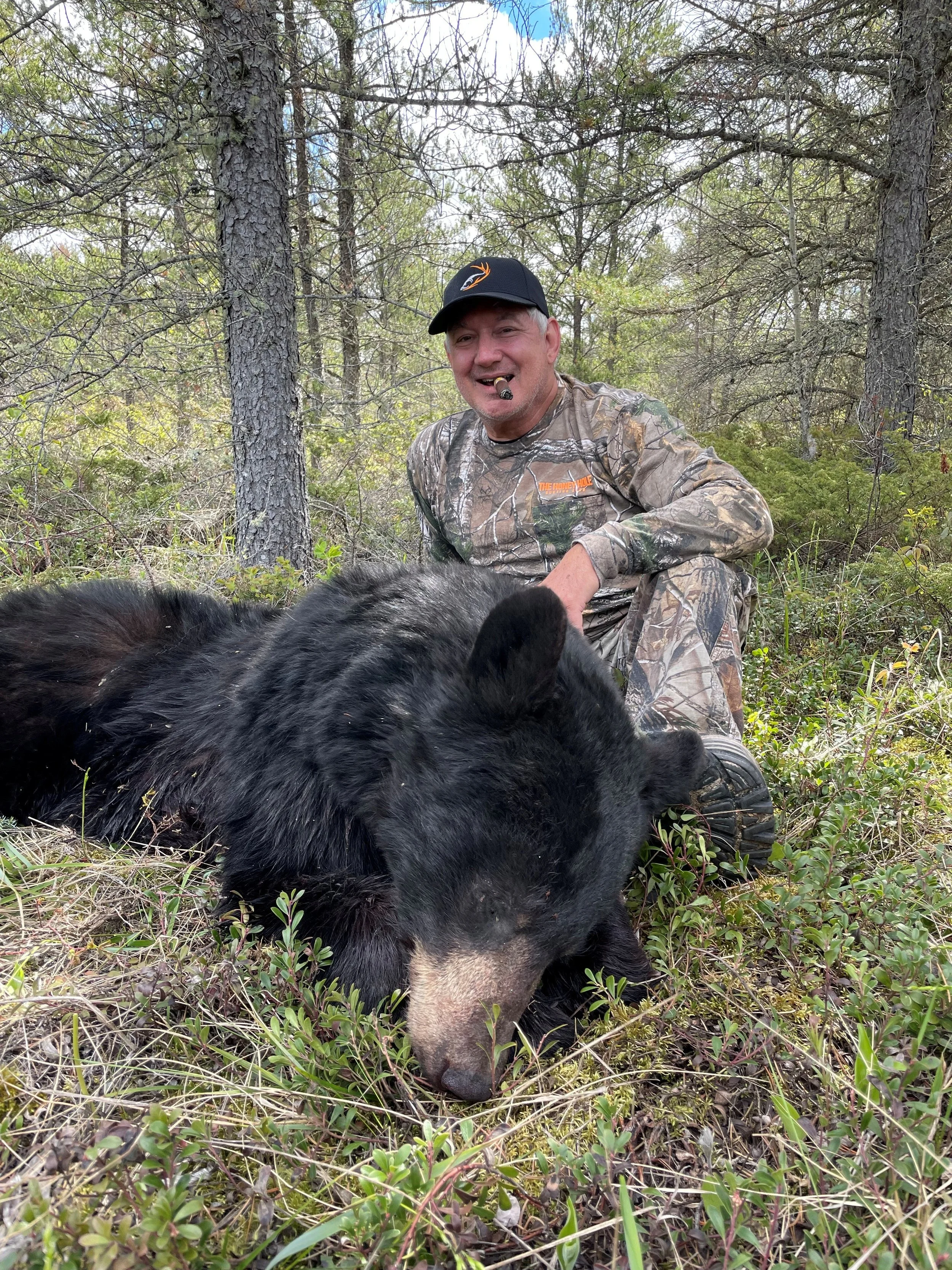 A man in camouflage sits next to a large black bear lying on the ground in a forested area, smiling with a cigar in his mouth.