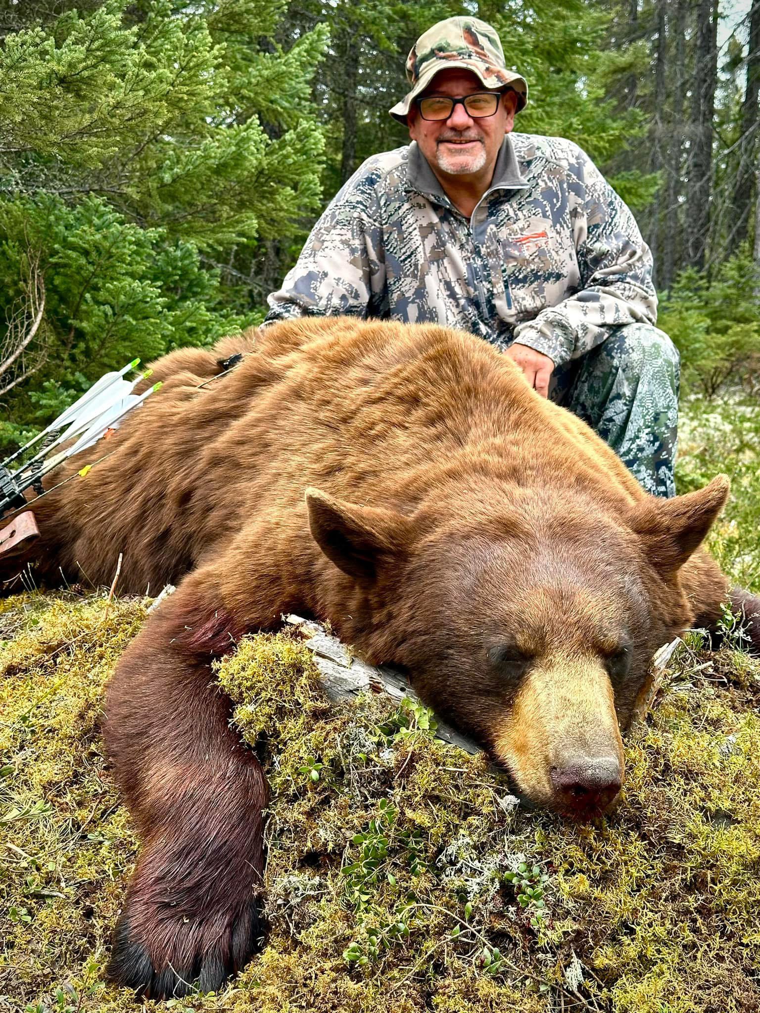 A man kneeling behind a large brown bear lying on mossy ground in a forest, with a bow and arrows visible on the bear's side.