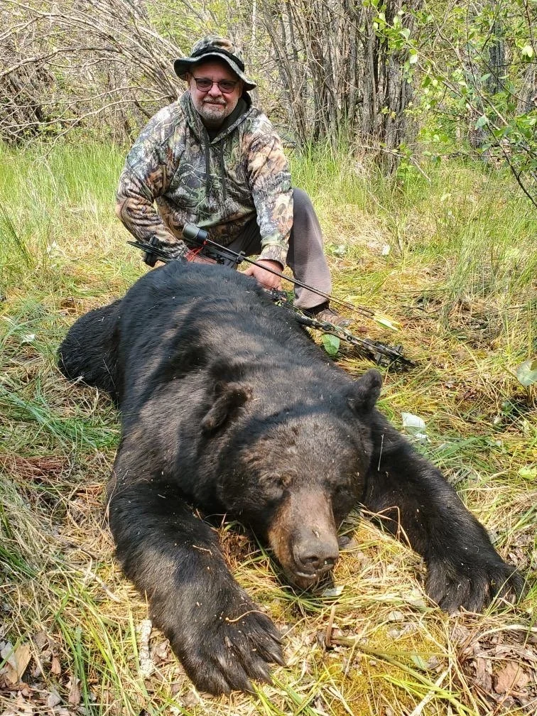 A man in camouflage and a wide-brimmed hat kneels next to a large, black bear lying on the ground in a forested area.