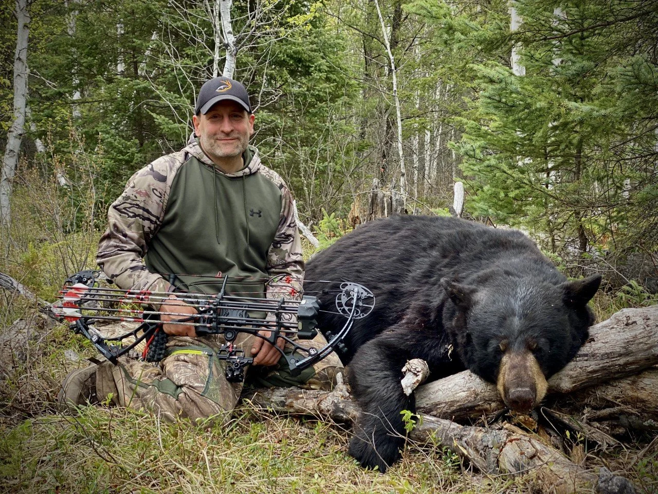 A man kneeling in a forest with a bow and arrows, next to a large, black bear lying on fallen logs.
