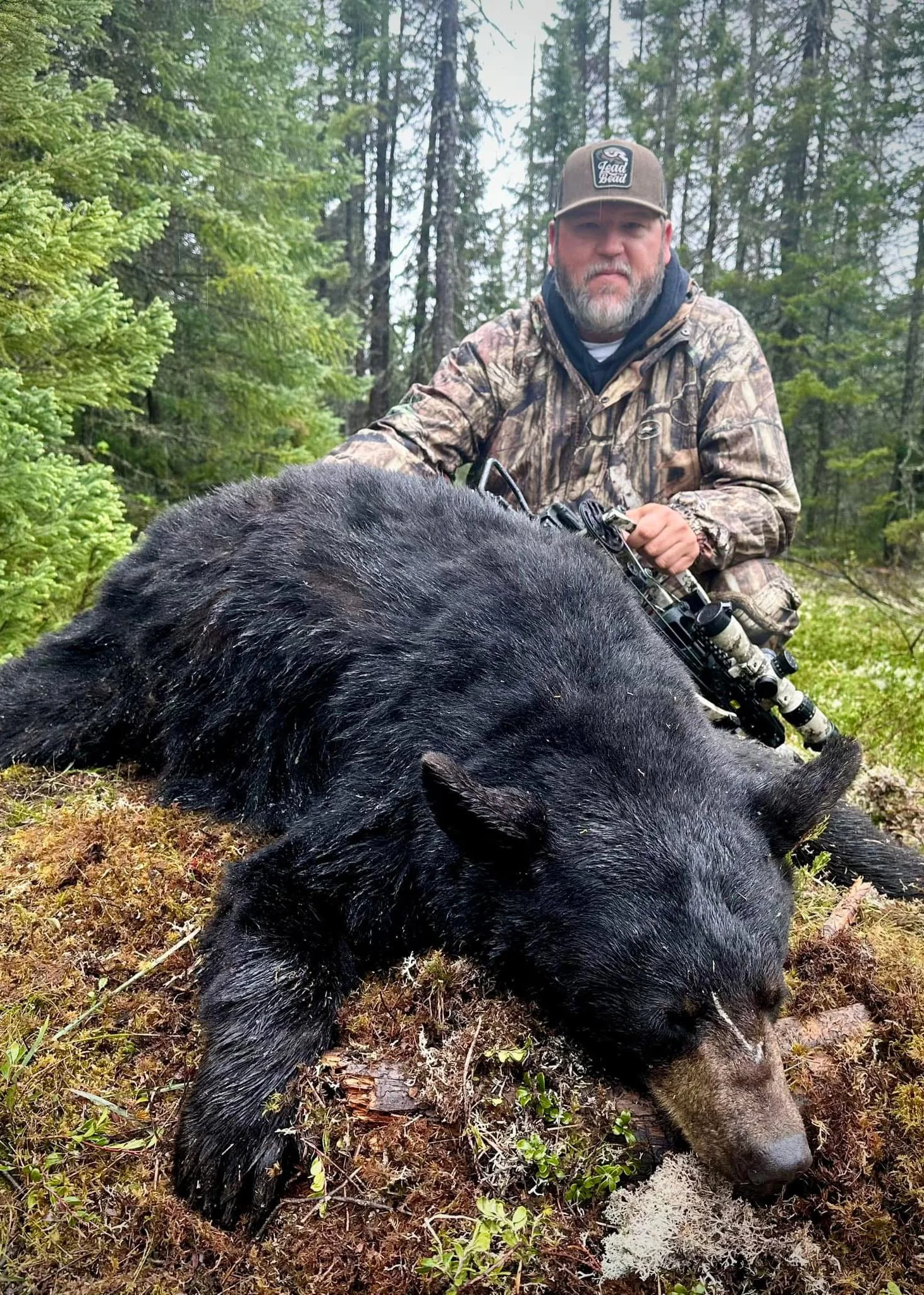 A bearded man wearing camouflage and a hat is kneeling in a forest beside a large black bear lying on the ground.