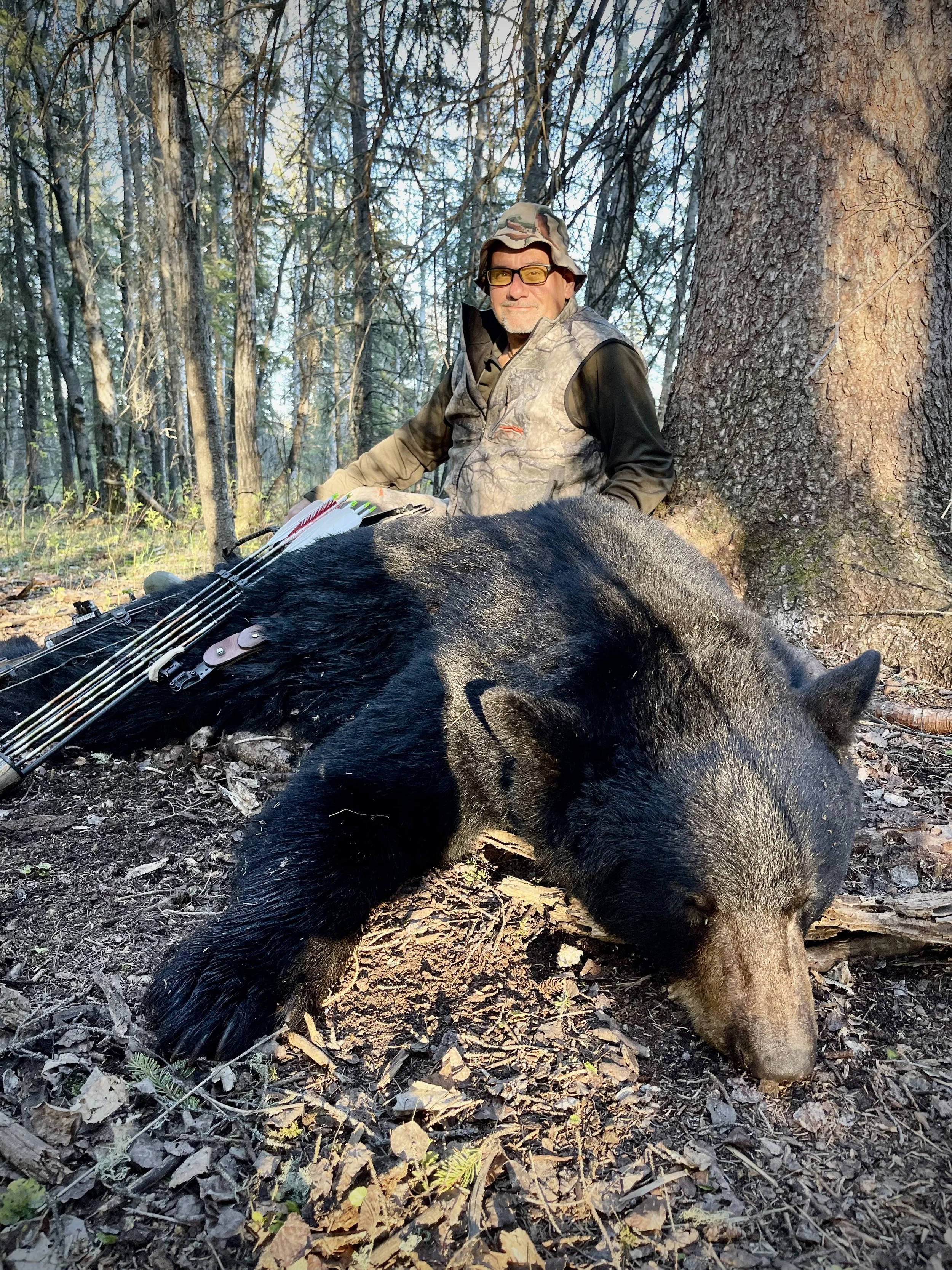 A man in camouflage poses next to a black bear in a forest, with a compound bow lying on the ground beside him.