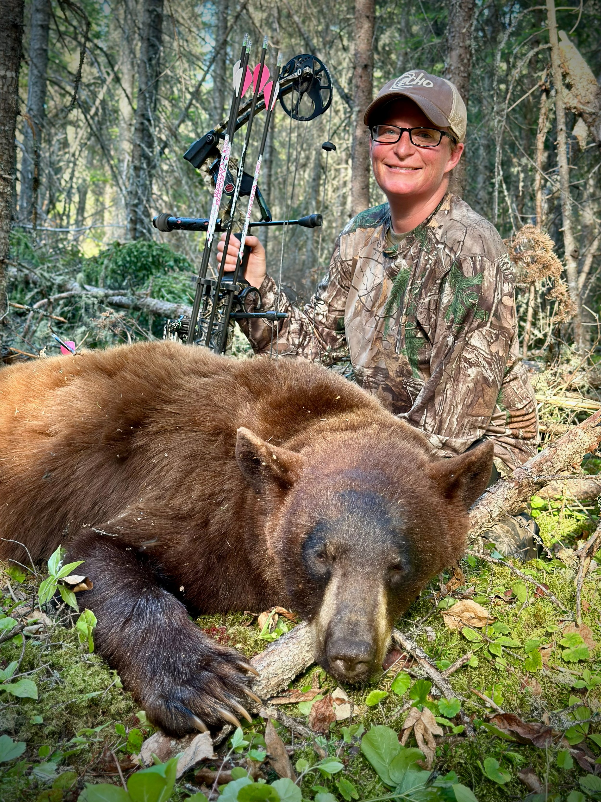 A woman in camouflage kneels next to a large, brown bear lying on the forest floor. The woman is smiling and holding a compound bow, with arrows in a quiver. The scene is set in a dense, green forest.