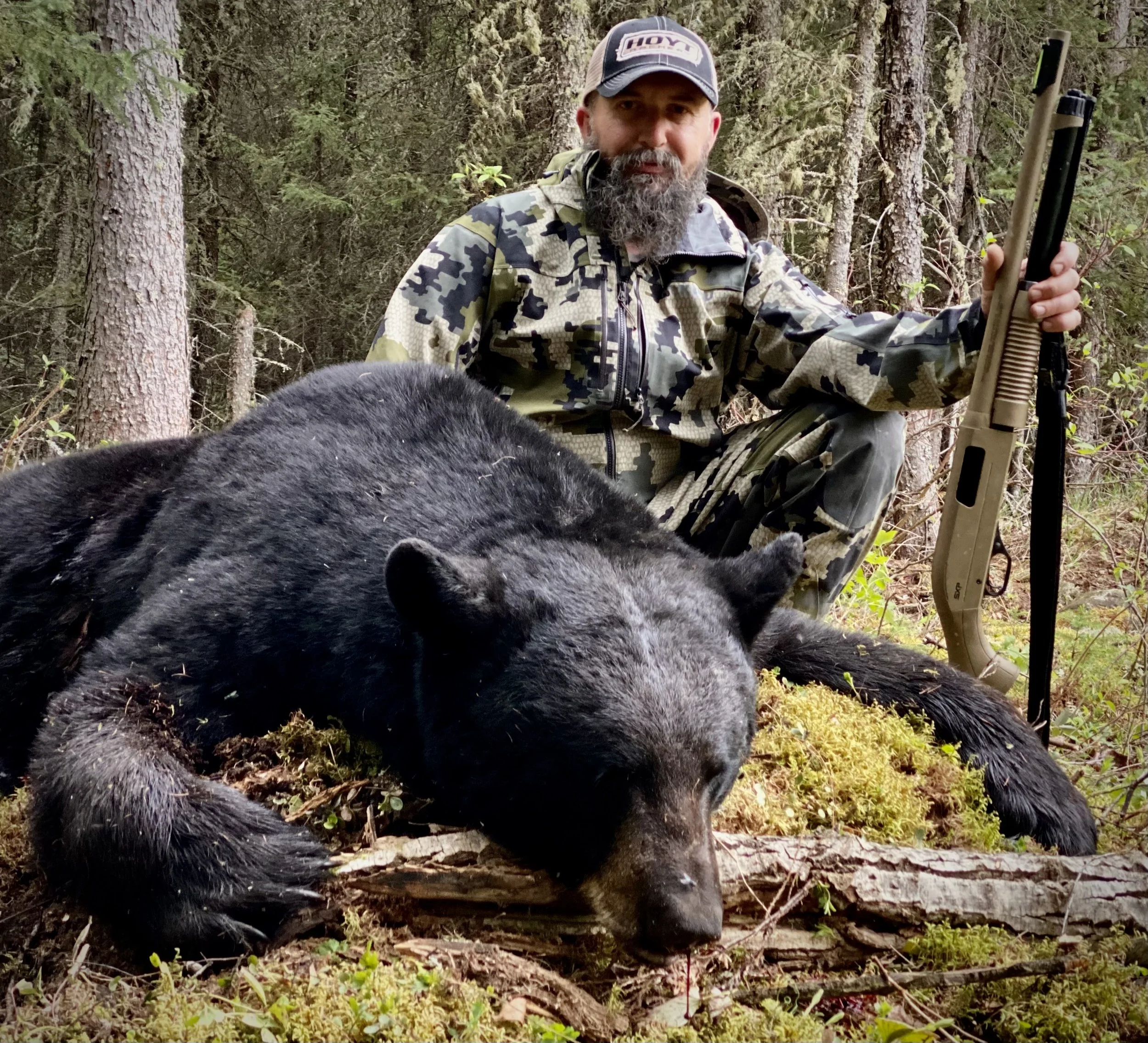 A man in camouflage crouches in a forest, holding a rifle, next to a large black bear lying on the ground.