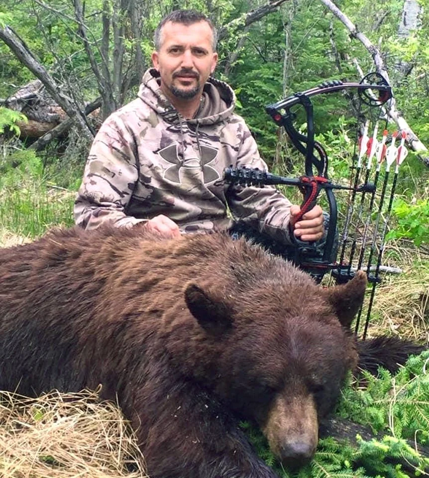 A man in a camouflage hoodie sits next to a large brown bear in a forest, holding a bow with arrows.