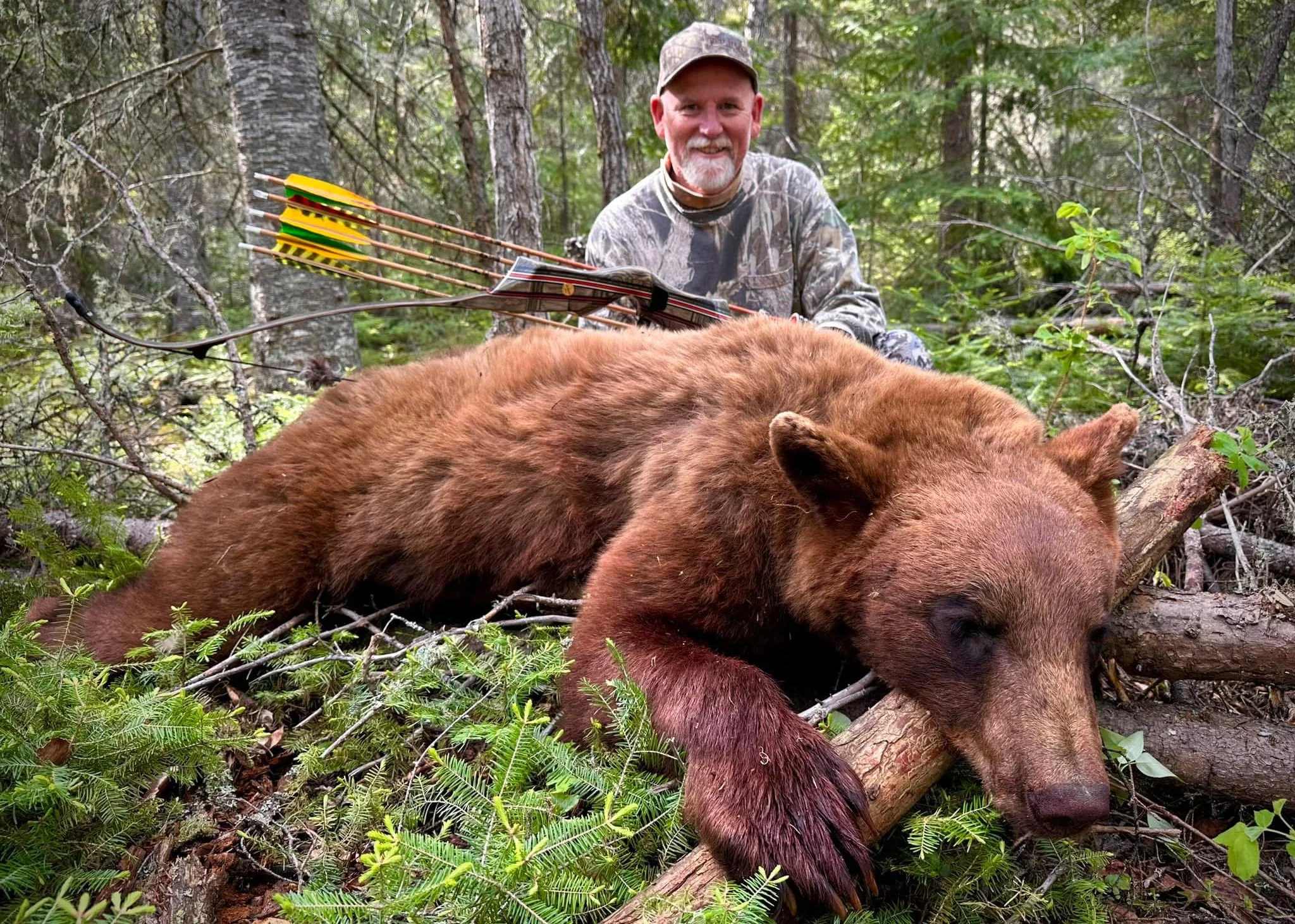 A man in camouflage is kneeling beside a large brown bear lying on the forest floor. The man is smiling and holding a bow with arrows in the background. The scene is set in a dense forest with green foliage and trees.