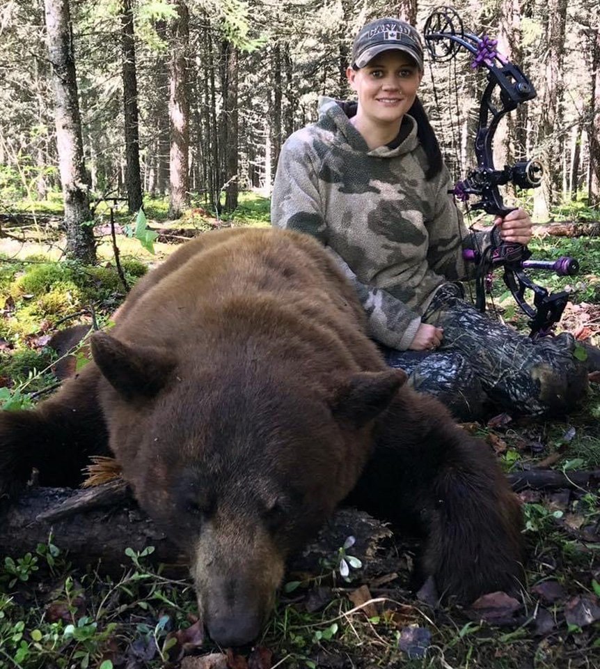 A woman in camouflage, holding a bow, is sitting in a forest next to a large brown bear.