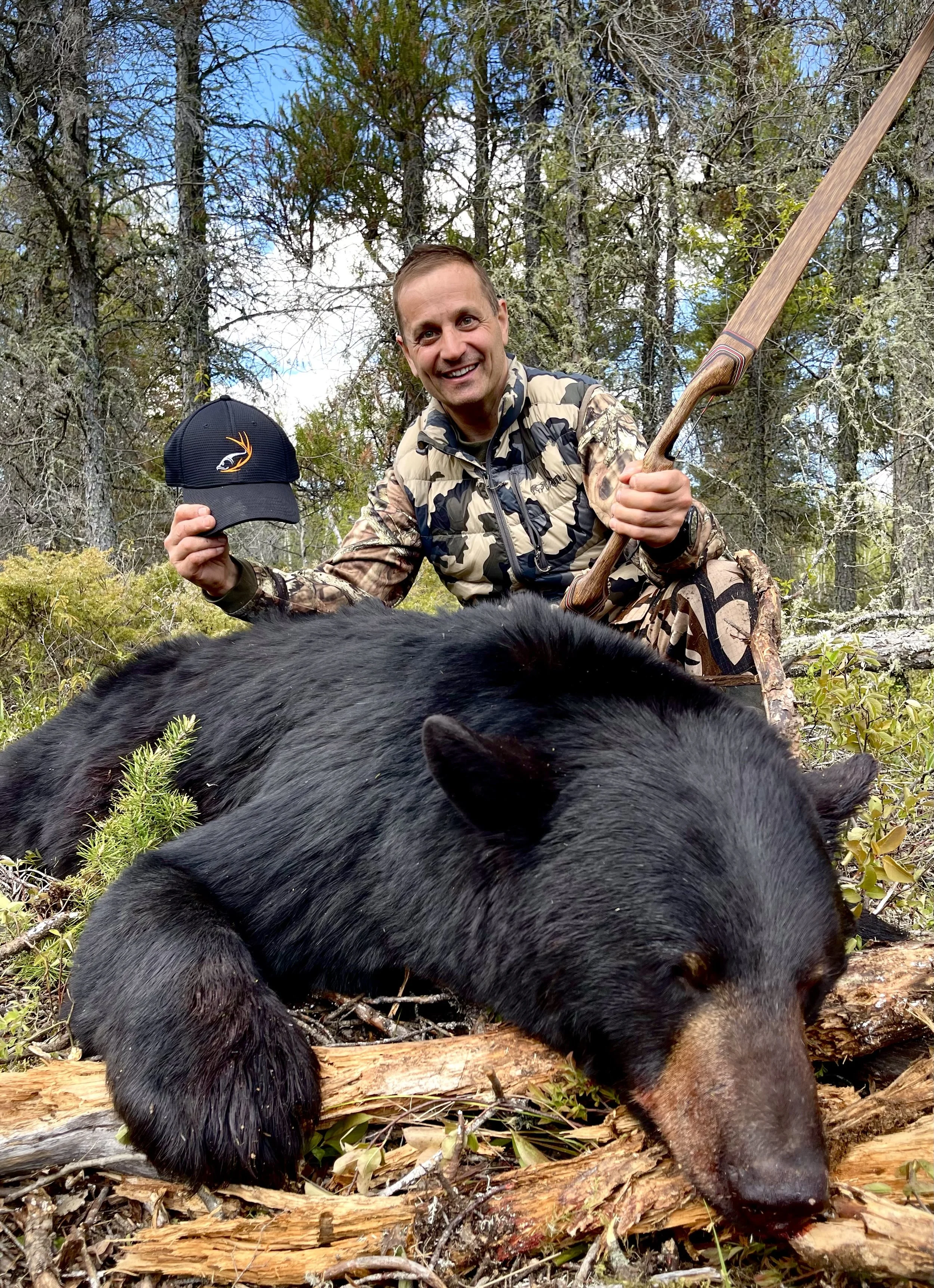 A man in camouflage clothing kneels in a forest, smiling as he holds a cap with the Target Species Outfitting logo, next to a black bear lying on the ground.