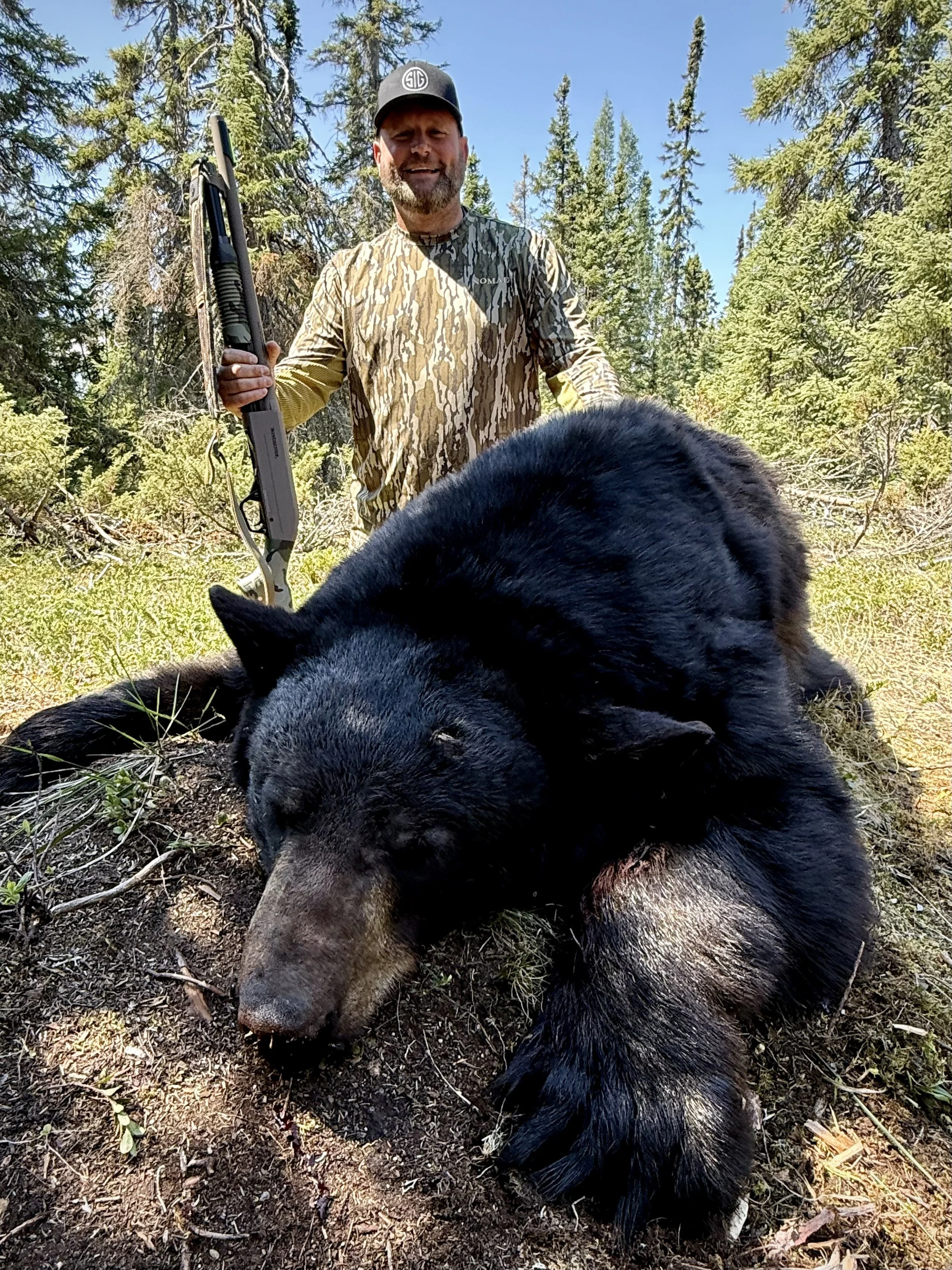 A man standing outdoors in a forested area, holding a shotgun, with a large, black bear lying on the ground in front of him.