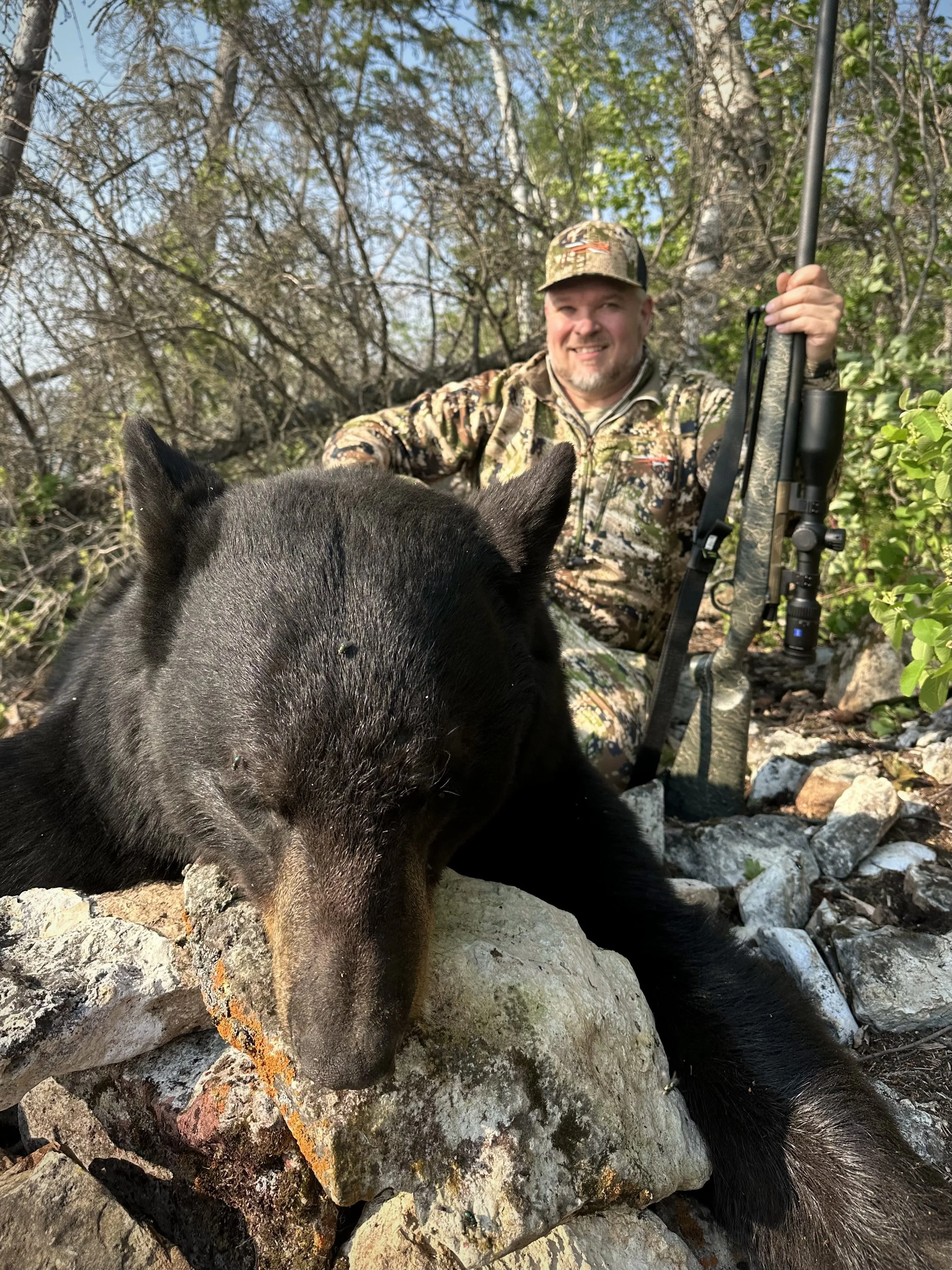 A man in camouflage is sitting on rocks in a wooded area, holding a rifle and smiling, with a black bear lying on a rock in front of him.