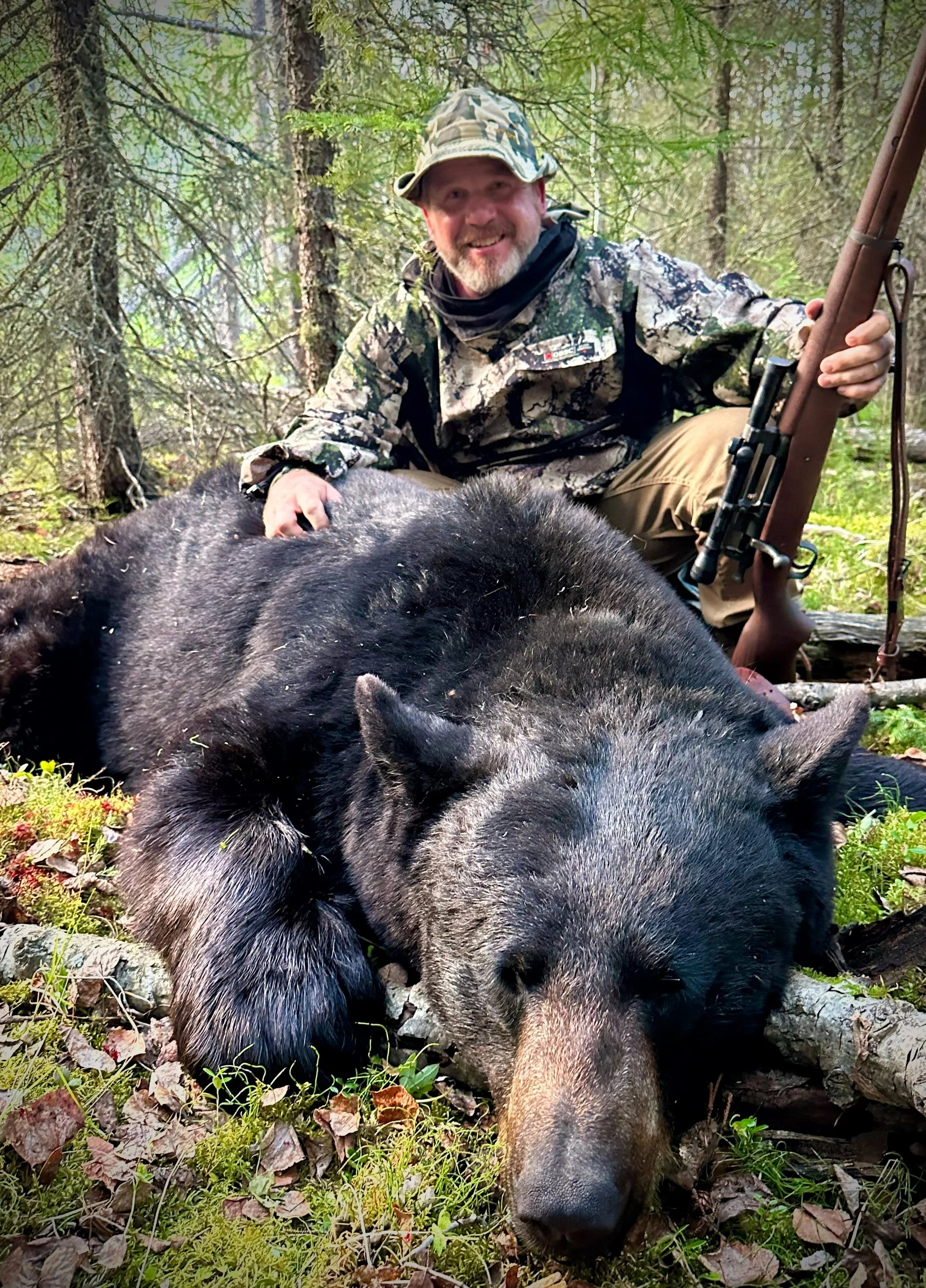 A man in camouflage is kneeling next to a large black bear lying on the ground in a forested area, holding a rifle and smiling.