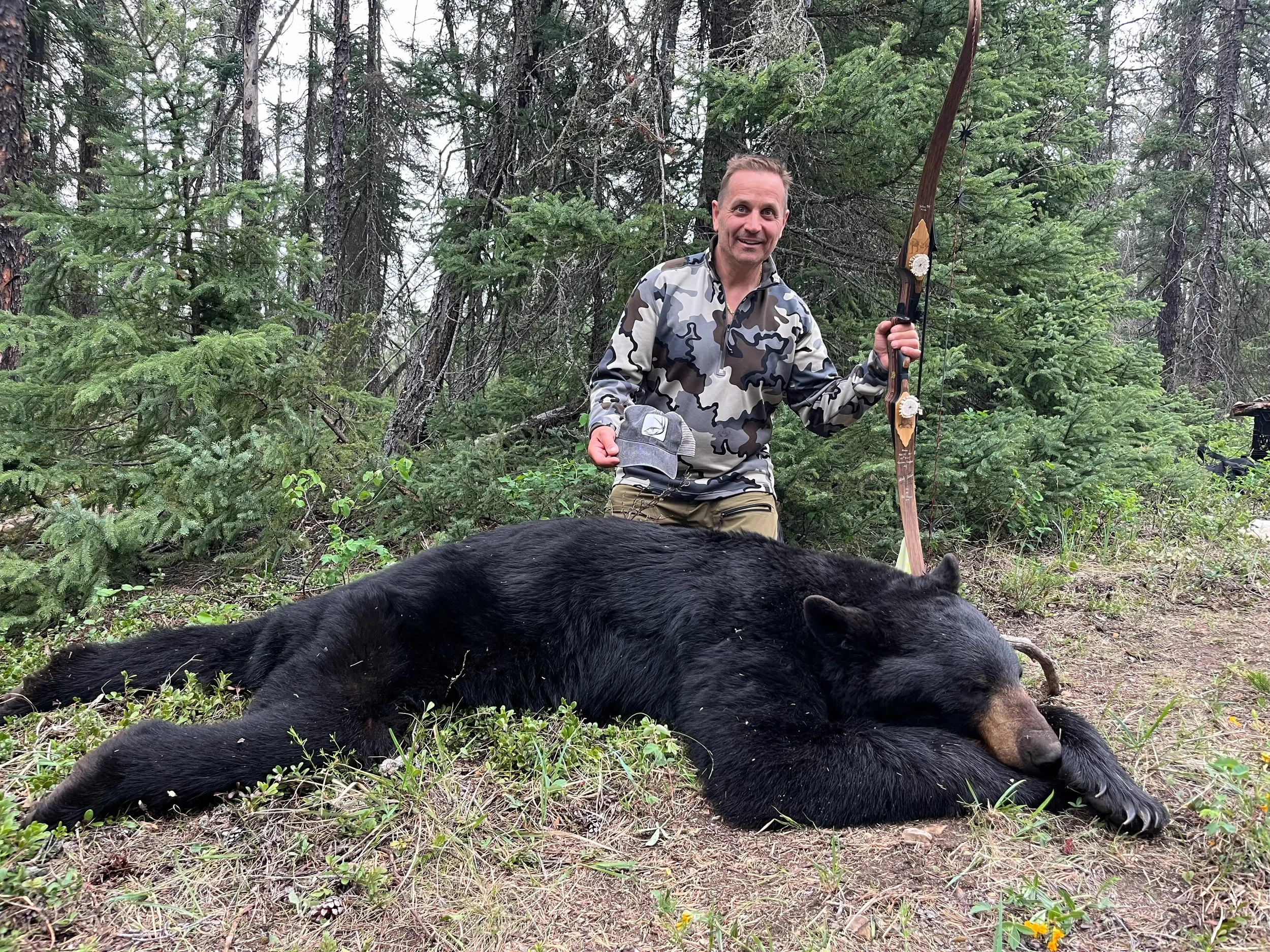 A man in camouflage is kneeling in a forest, holding a bow, next to a large black bear lying on the ground. The man is smiling, and the forest background is filled with trees and green foliage.