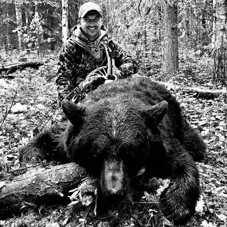 A smiling man in camouflage and a hat kneels next to a large black bear in a forest.